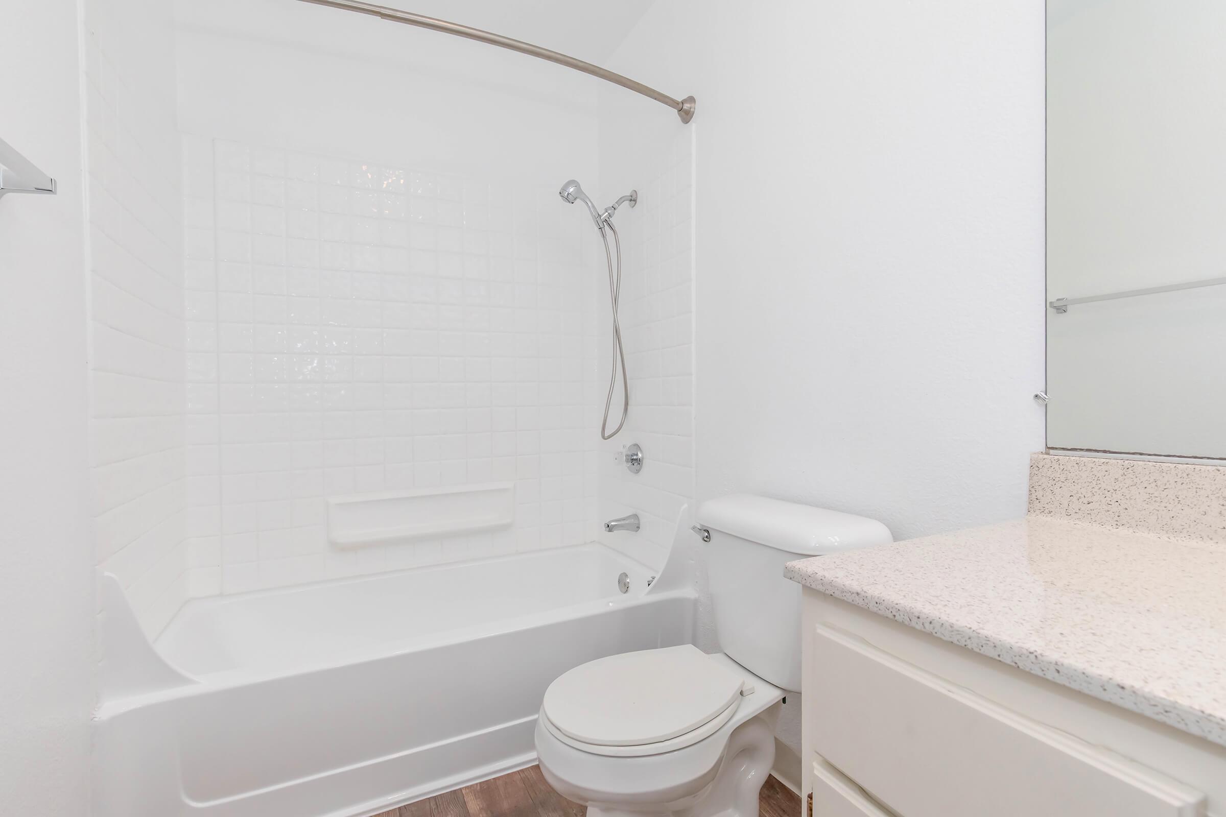 A clean, modern bathroom featuring a bathtub with a shower attachment, a white toilet, and a vanity with a granite countertop. The walls are painted white, and the floor is wood-like. A mirror is mounted above the vanity, creating a bright and airy atmosphere.