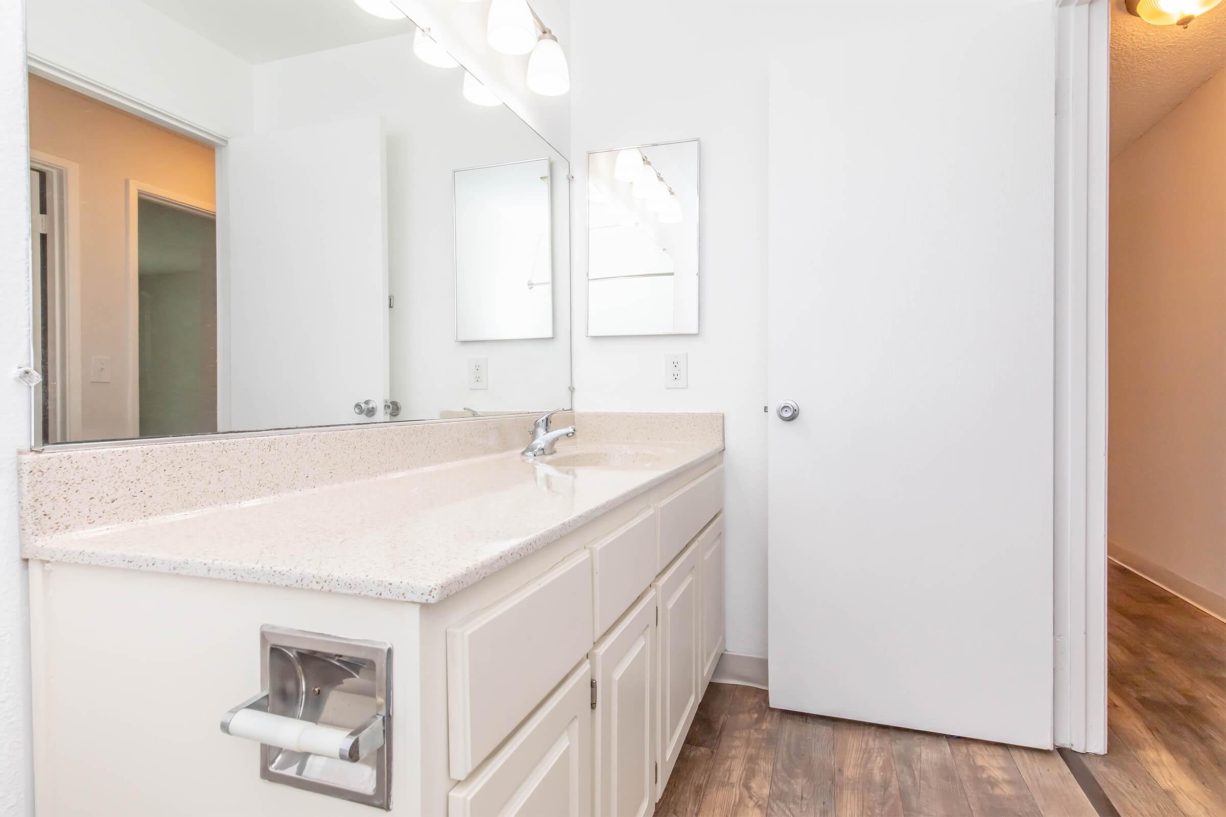 A modern bathroom with a spacious vanity featuring a light-colored countertop and two mirrors above. The walls are white, and the flooring is a light wood finish. A door leads to another area, while a window provides natural light. Bright lighting fixtures are mounted above the mirrors.