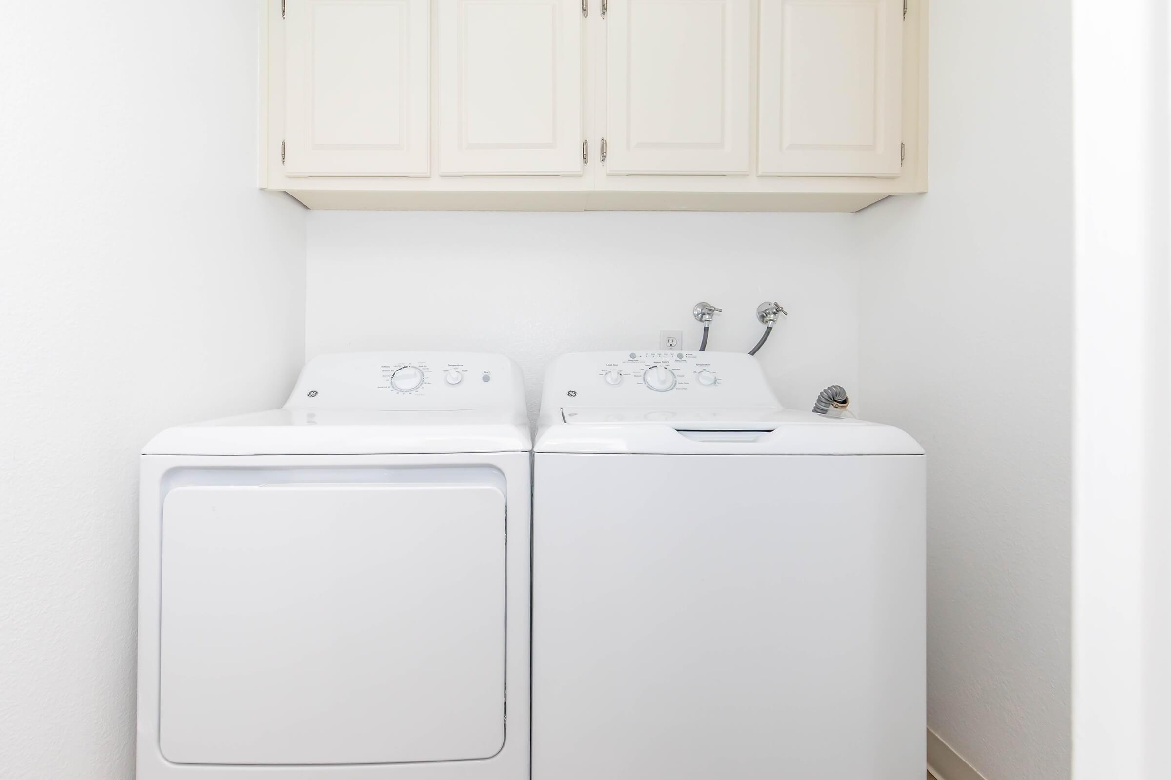 A laundry area featuring a white washing machine and dryer side by side, with a light-colored cabinet above. The walls are painted white, creating a clean and bright space.