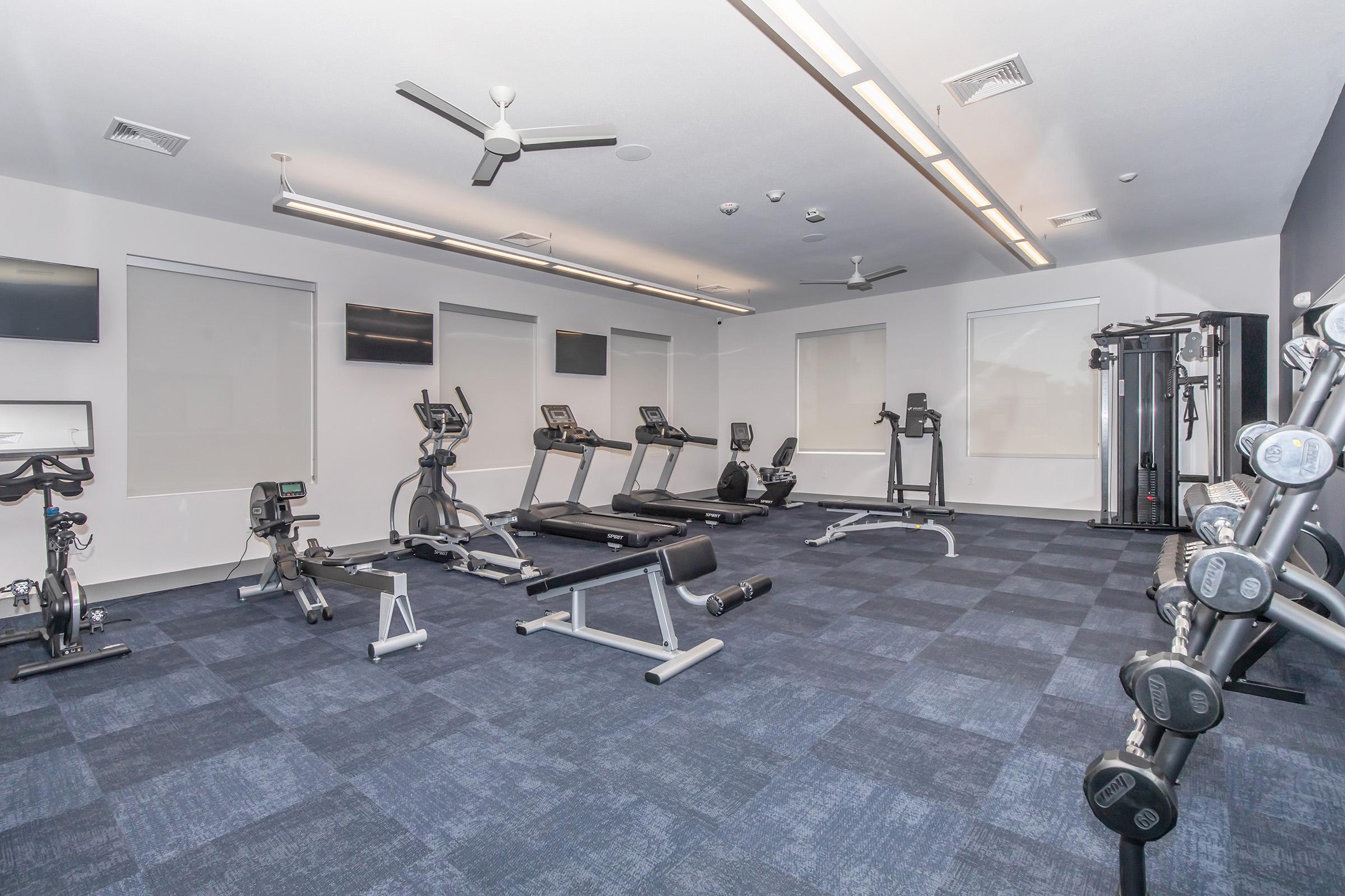 A modern gym interior featuring various exercise equipment, including treadmills, elliptical machines, weight benches, and free weights. The space has a blue carpet, ceiling fans, and large windows with natural light. There are also wall-mounted screens for entertainment or instruction.
