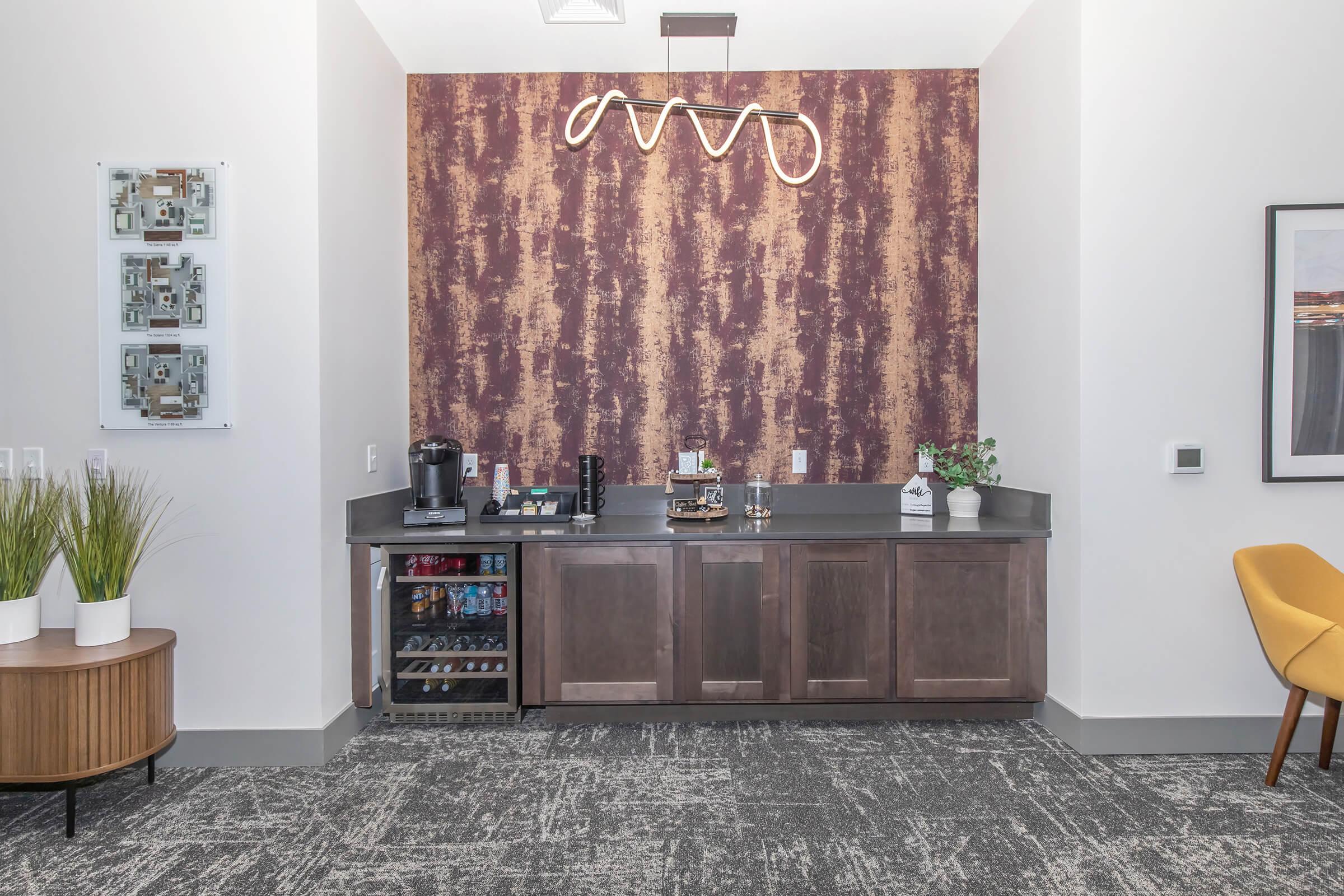 A modern kitchenette area with a dark countertop and wooden cabinets. The wall behind features a textured, patterned wallpaper in warm tones. On the counter, there’s a coffee maker, a small decorative plant, and a drink fridge. A yellow chair is positioned nearby, and there are light fixtures above for illumination.