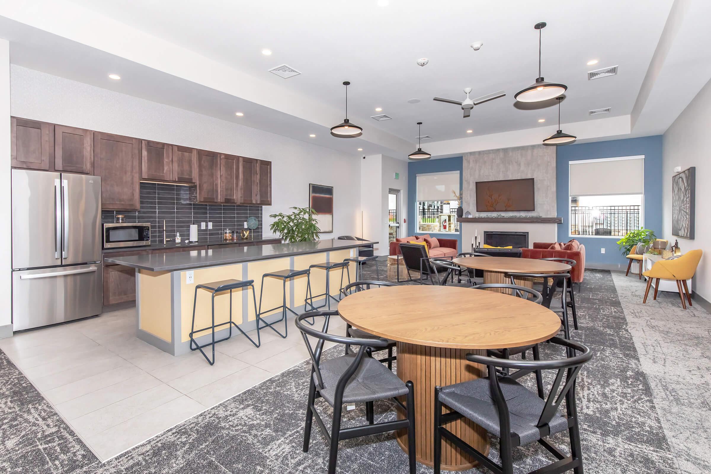 Modern community lounge with a kitchen area featuring dark wood cabinetry and stainless steel appliances. There are several tables, including round and rectangular options, surrounded by black chairs. Comfortable seating areas with stylish chairs are positioned nearby, along with a decorative plant and a fireplace.