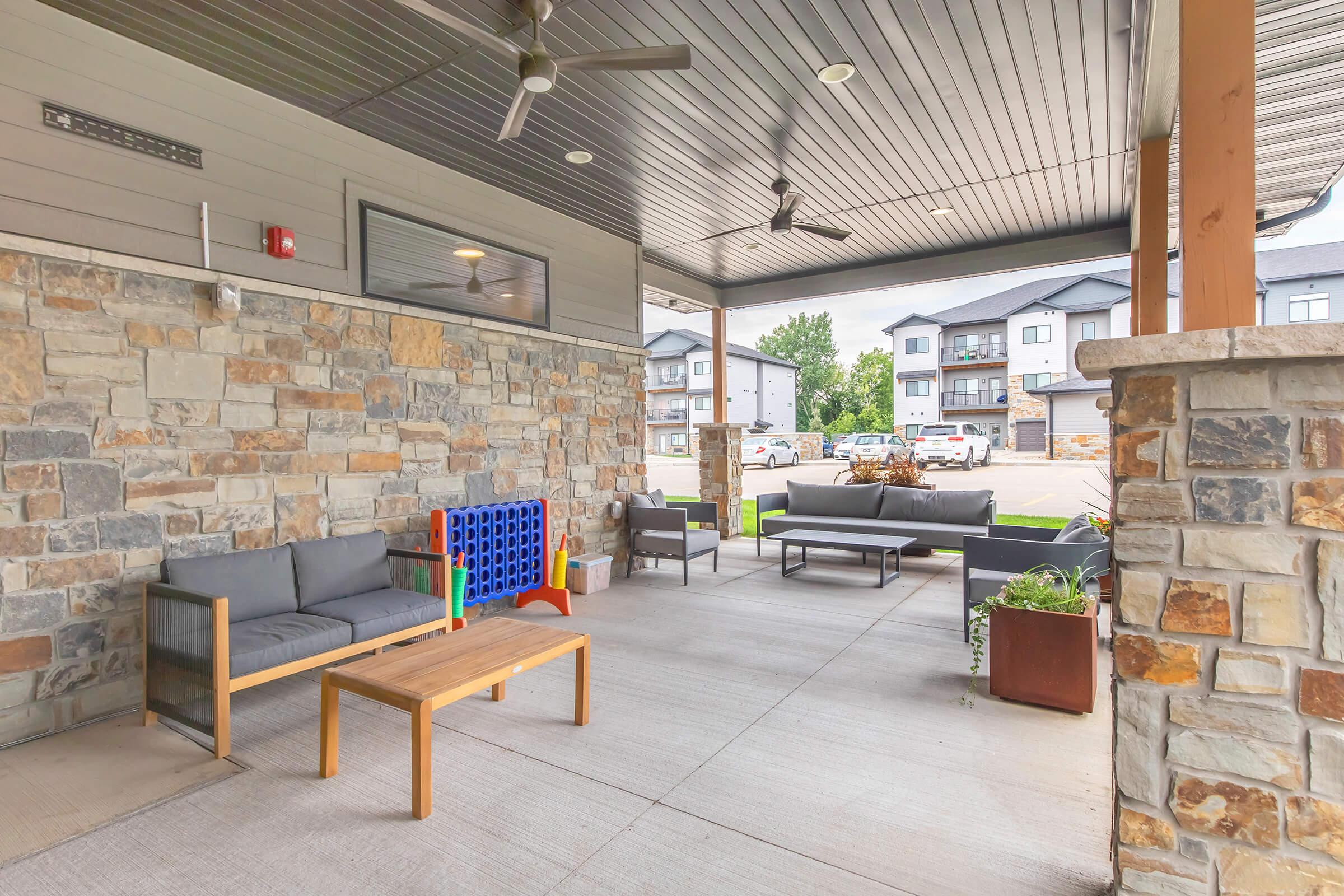 A covered outdoor seating area featuring a gray couch, wooden coffee table, and colorful children's play structure. The space is surrounded by stone walls and has a modern design, with nearby parking visible in the background and plants in decorative pots.