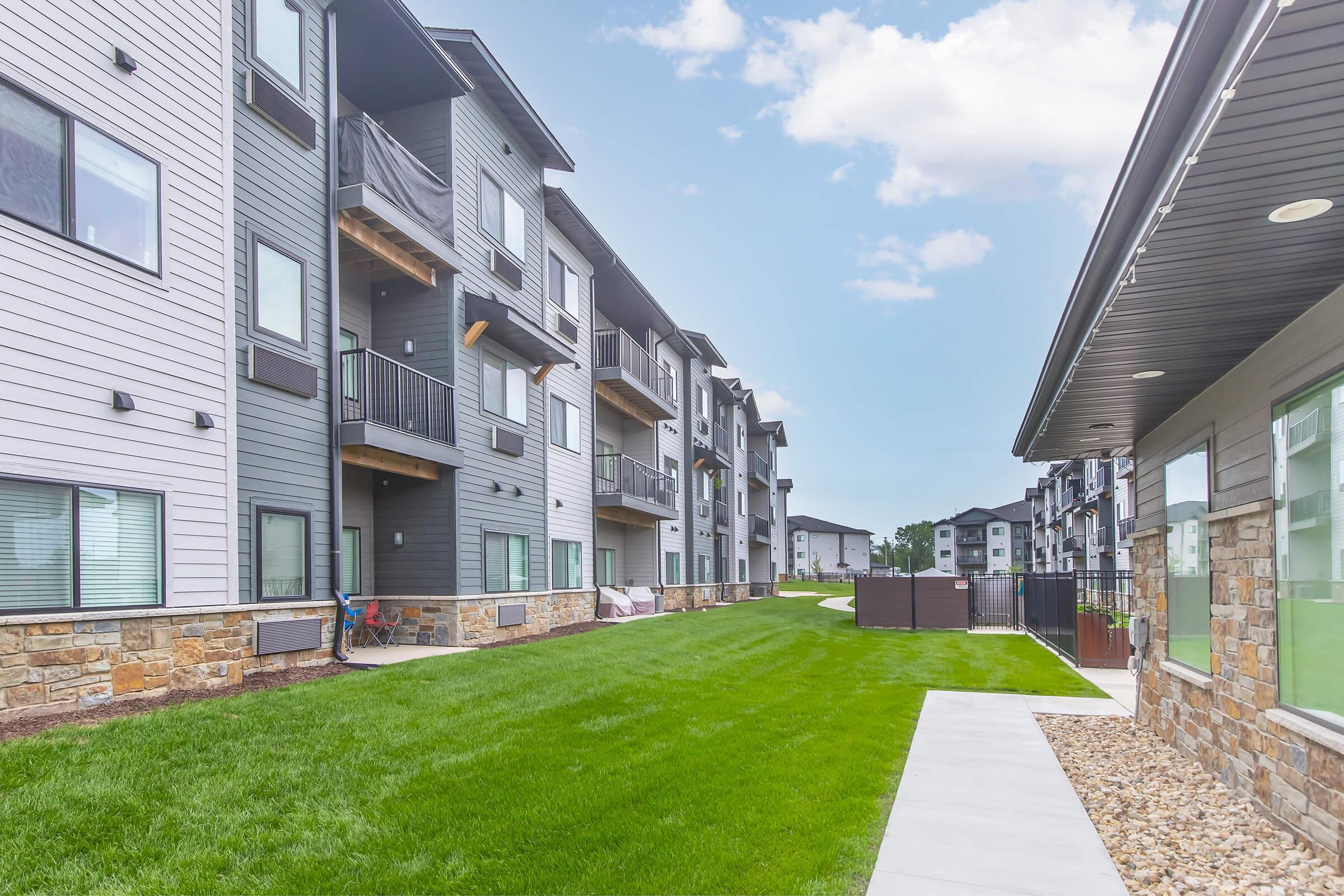 A view of a modern apartment complex with multiple three-story buildings. The buildings feature a mix of gray siding and stone accents, with balconies on the upper floors. A well-maintained green lawn runs alongside a concrete walkway, and there are fences and outdoor amenities visible in the background.