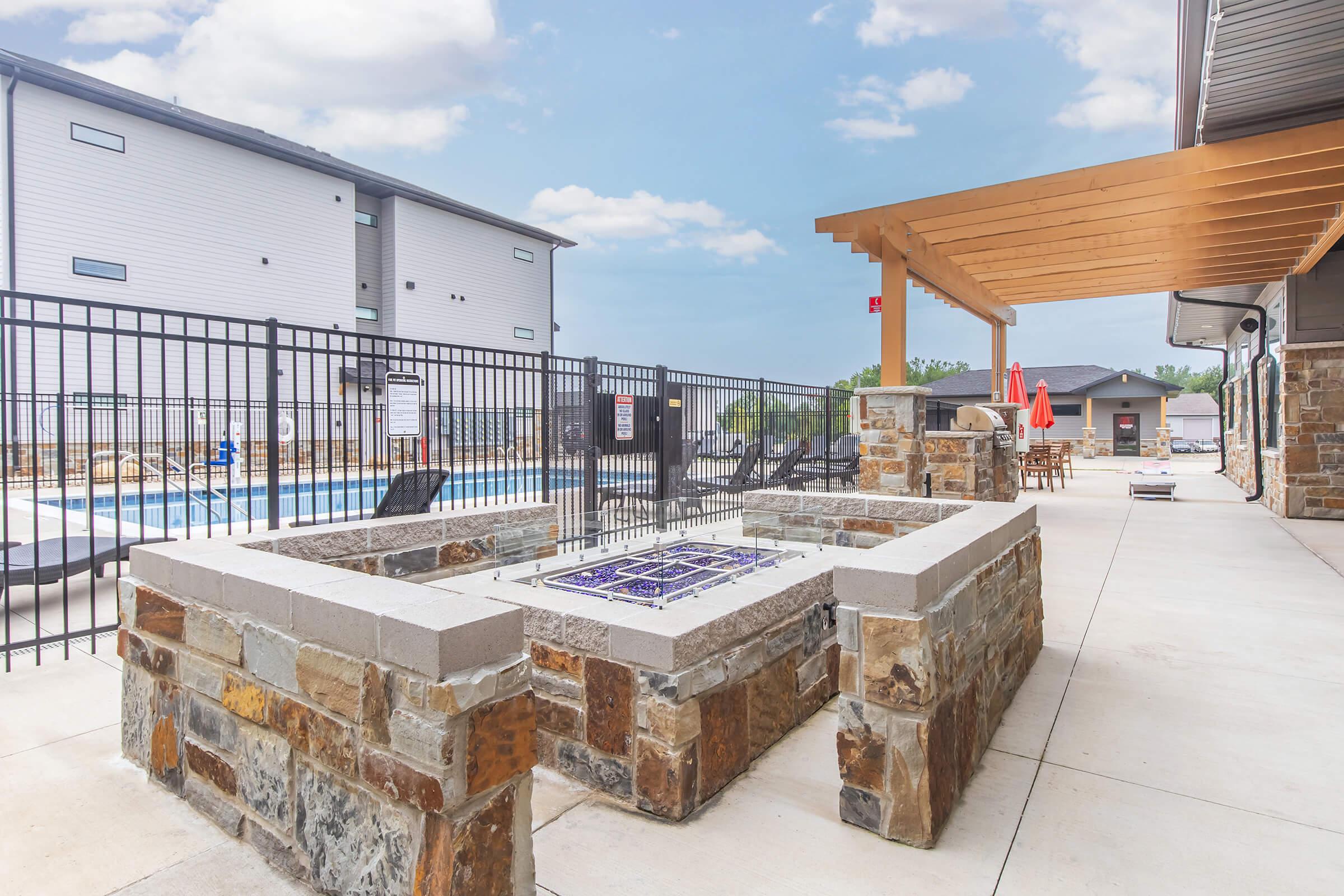 Outdoor area featuring a stone fire pit with decorative tiles, surrounded by a fenced space. In the background, a swimming pool is visible, along with lounge chairs and umbrellas. A covered seating area is present nearby, with tables and chairs under a wooden pergola. Bright blue sky with scattered clouds above.