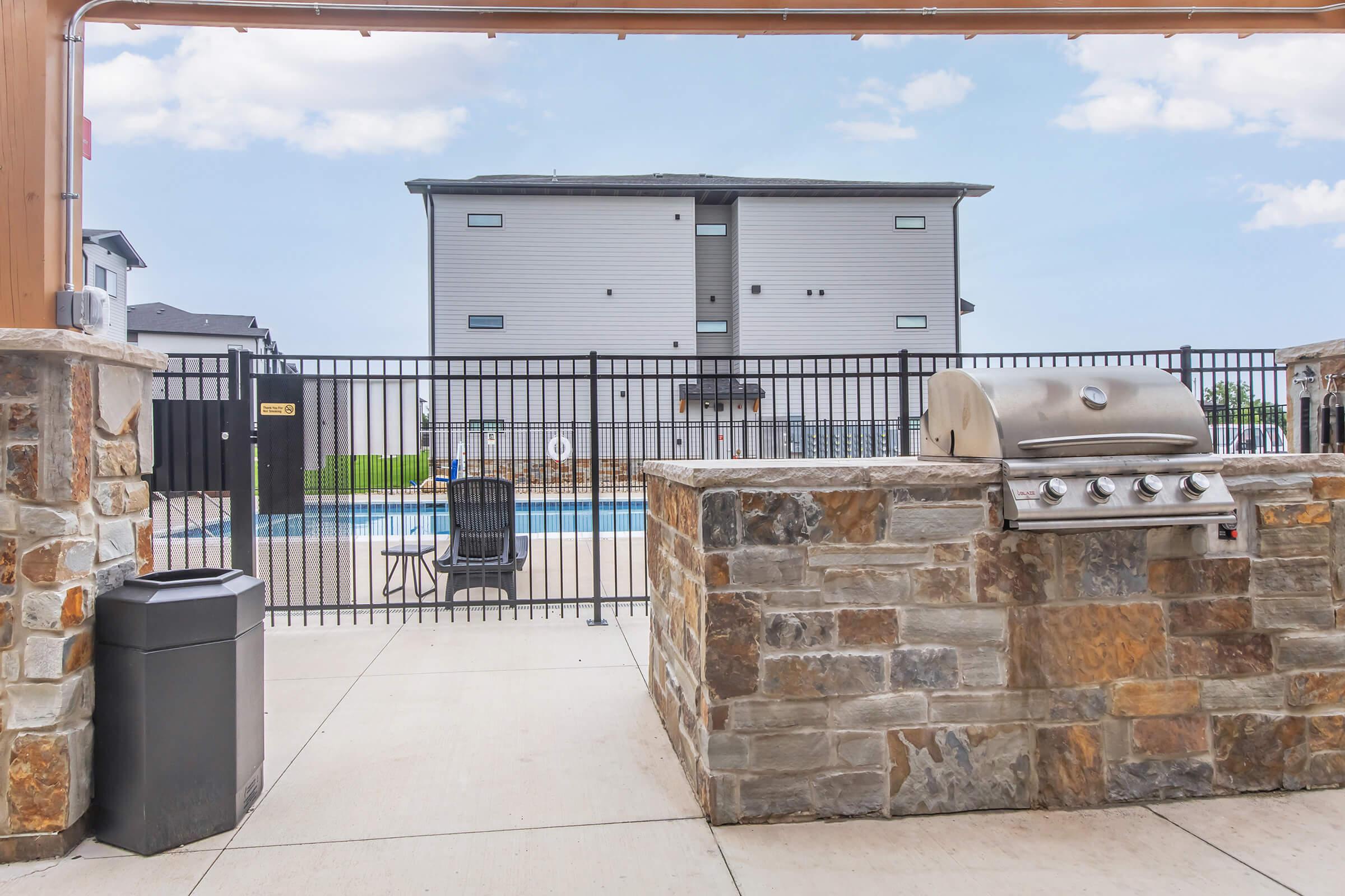 Outdoor pool area featuring a fenced-in swimming pool in the background. In the foreground, there is a stone barbecue grill and a trash can. A single chair is positioned near the pool, with a modern multi-story building visible beyond the fence. The sky is clear and bright.