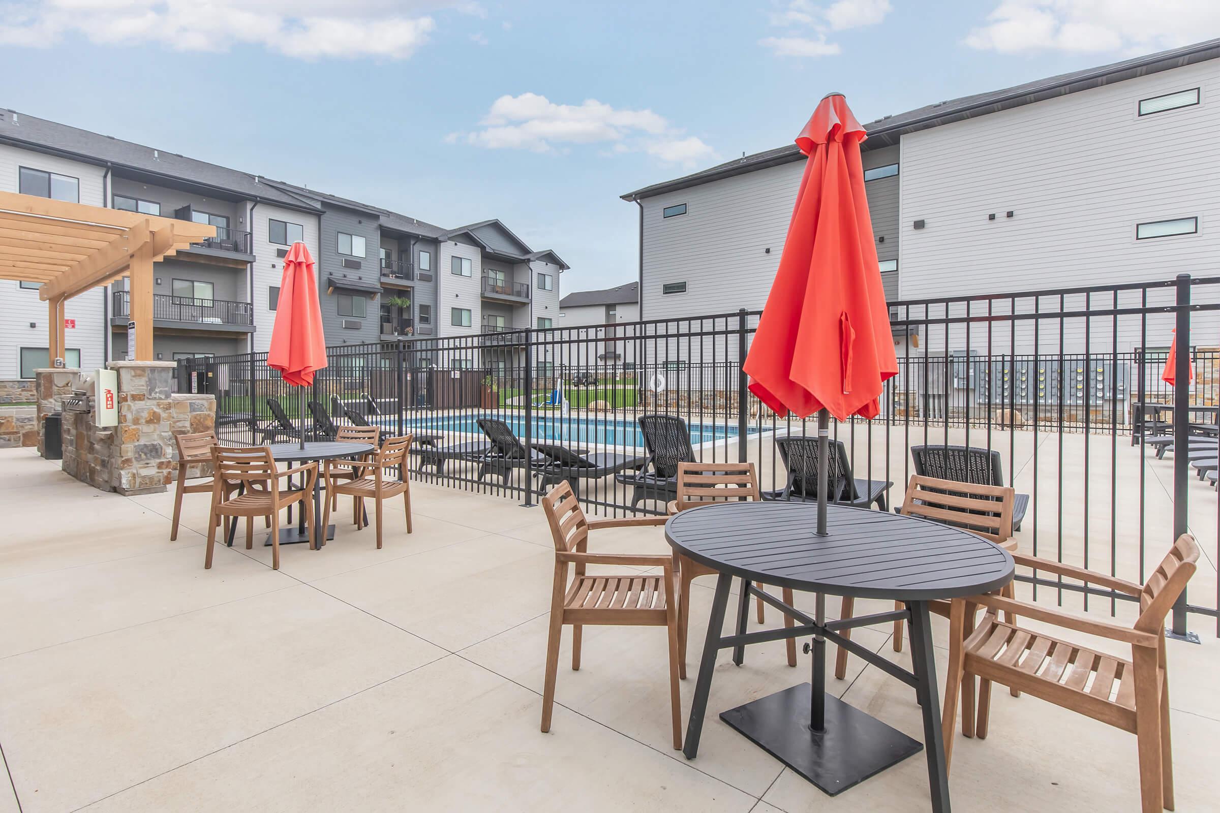 Poolside area featuring wooden tables and chairs with red umbrellas, surrounded by a black fence. In the background, a swimming pool is visible, along with buildings that have modern architecture. The scene is set under a clear blue sky, creating a relaxing atmosphere.