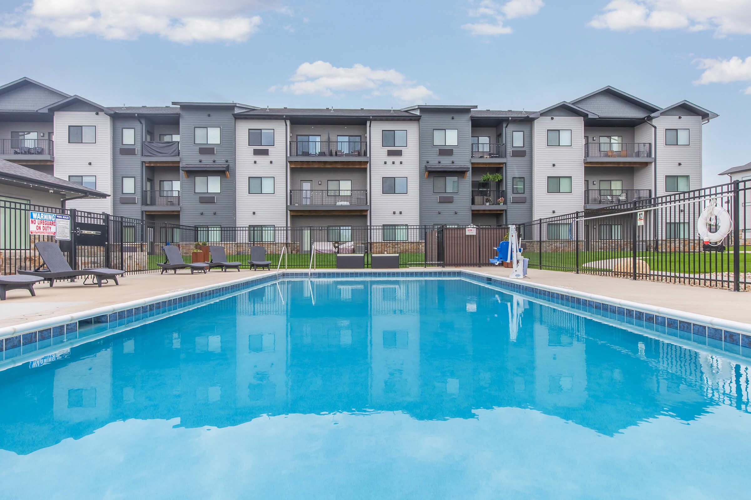 Swimming pool in front of an apartment complex. The pool is surrounded by a black fence and lounge chairs, with the building visible in the background. The sky is clear with a few clouds, reflecting on the surface of the water.