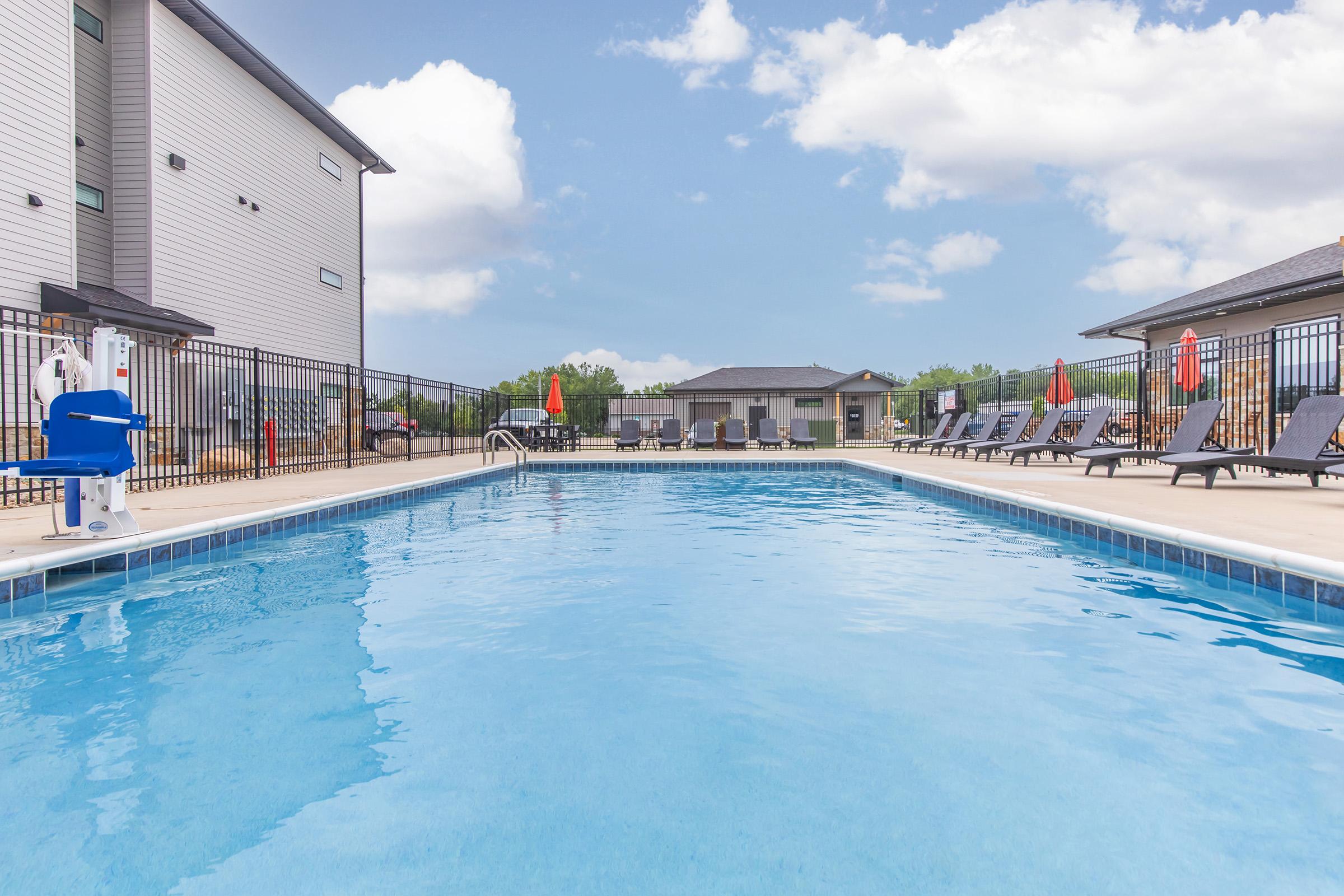A serene outdoor swimming pool surrounded by lounge chairs and umbrellas, with a clear blue sky and fluffy clouds overhead. The pool area is enclosed by a black fence, and adjacent buildings can be seen in the background, creating a relaxing atmosphere for leisure and swimming.
