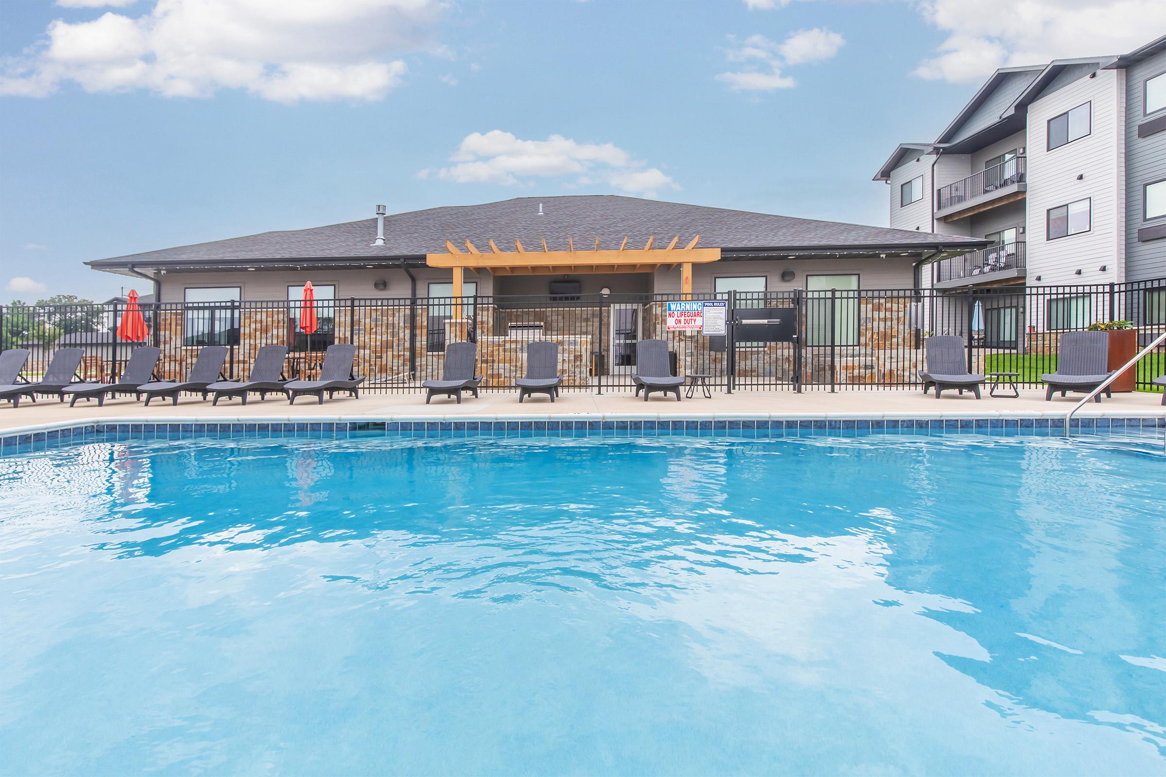 A clear, inviting swimming pool with lounge chairs lined up along the edge. In the background, there is a building with a modern design, featuring large windows and a decorative wooden canopy. The sky is partly cloudy, enhancing the bright, cheerful atmosphere of the pool area.