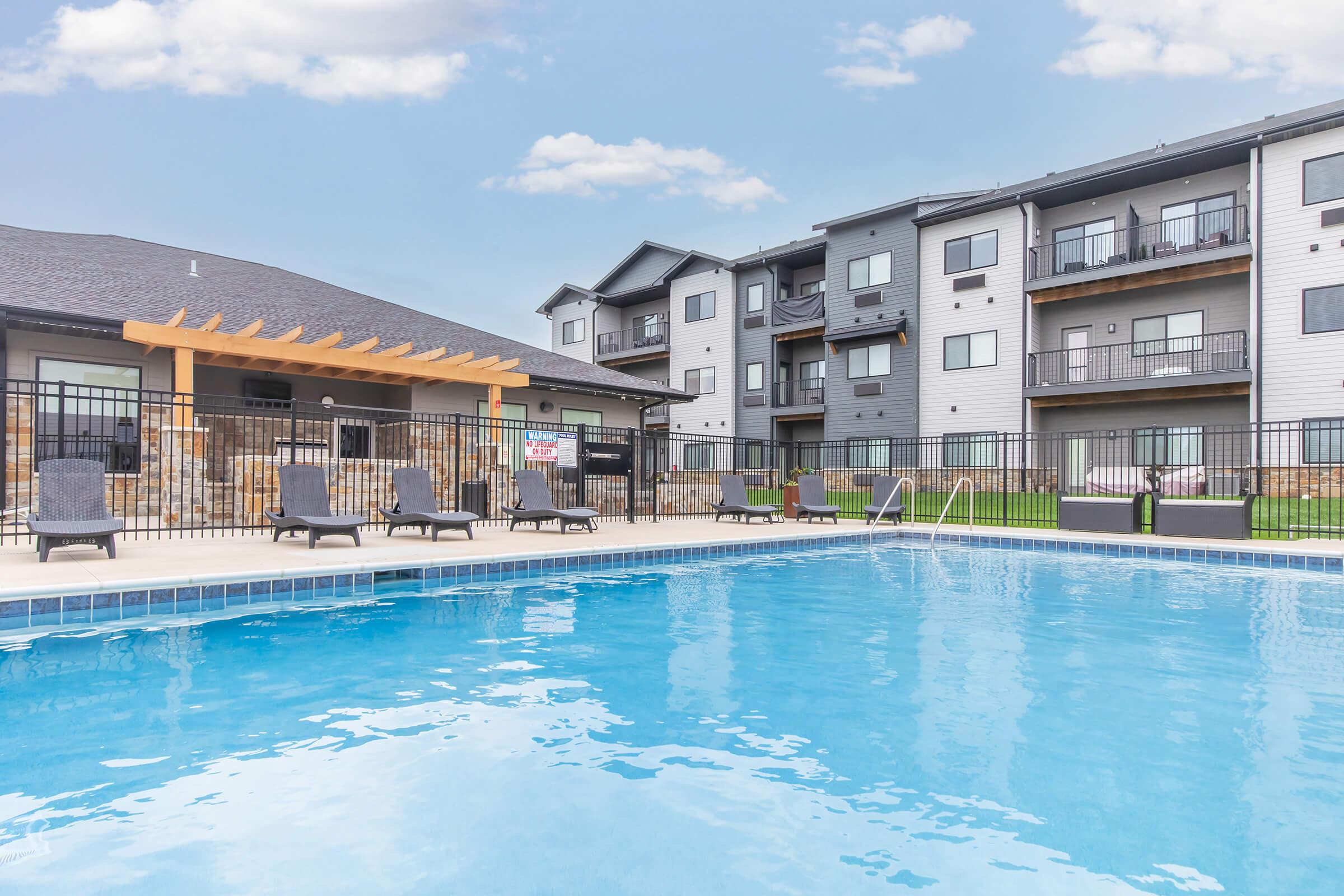 A clear swimming pool with lounge chairs arranged around it, surrounded by a fence. In the background, a multi-story residential building features balconies. The sky is blue with a few clouds, creating a peaceful atmosphere.