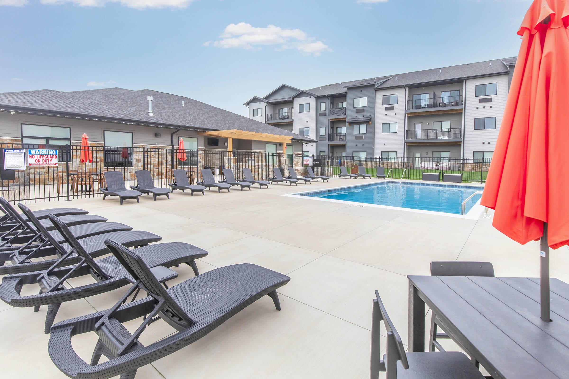 A spacious outdoor pool area featuring a clear blue swimming pool surrounded by lounge chairs and tables. Red umbrellas provide shade, and there are modern apartment buildings in the background under a bright blue sky. The scene conveys a relaxing atmosphere ideal for leisure and enjoyment.
