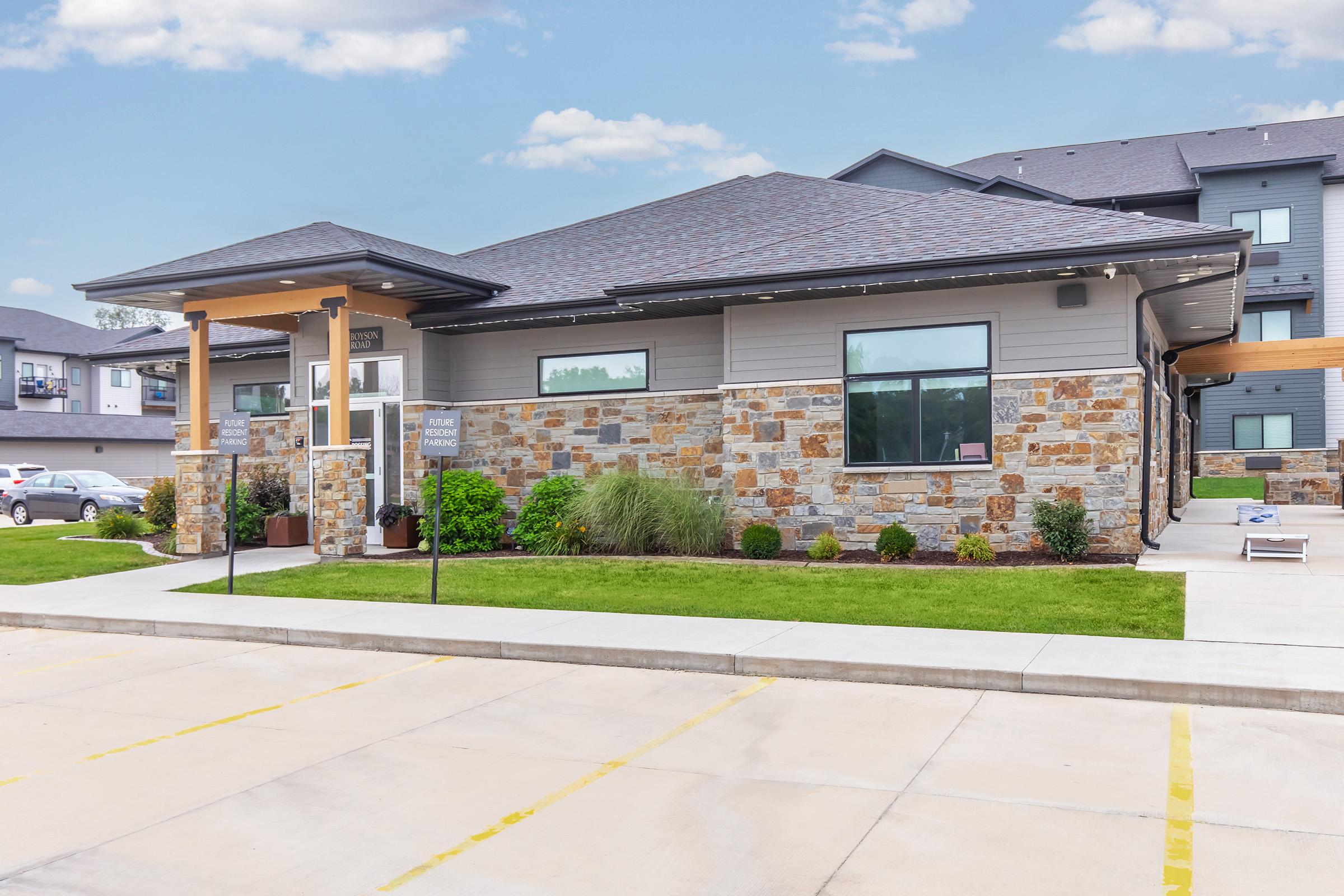 A modern building with a stone and siding exterior, featuring large windows and landscaping. The entrance has a covered porch with a sign. Parking spaces are visible in the foreground, and additional buildings can be seen in the background under a clear sky.