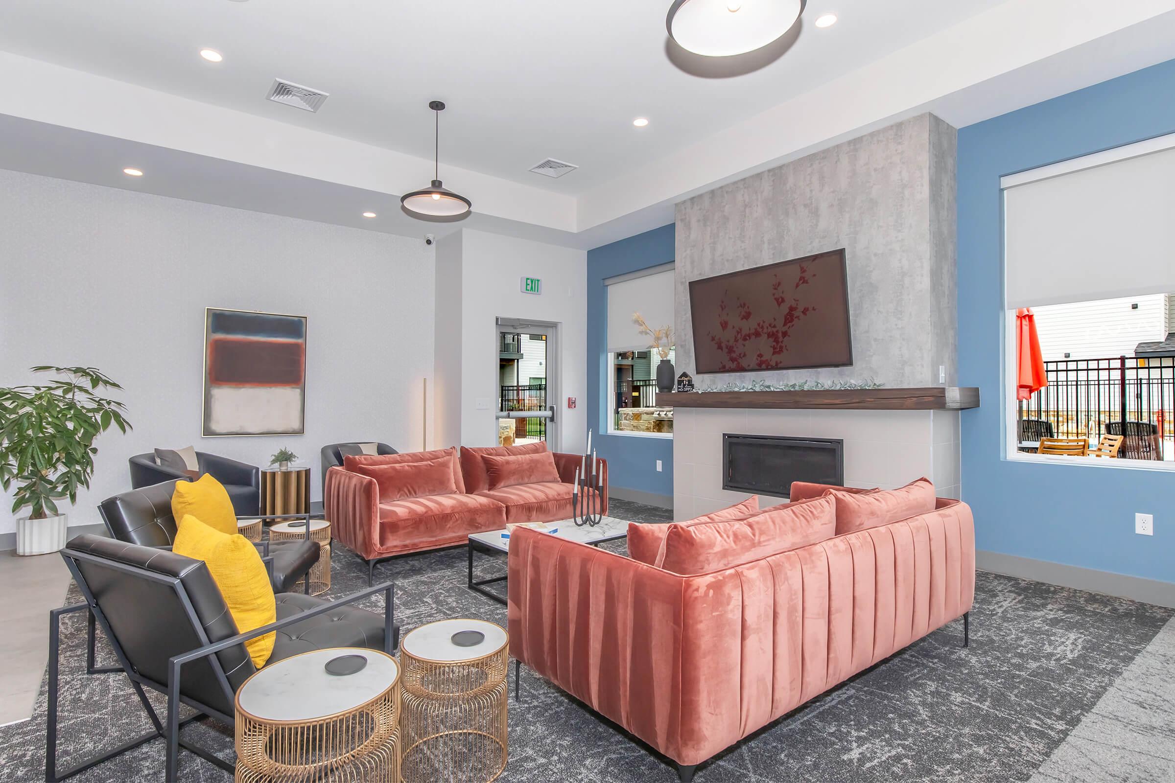 A modern lounge area featuring two pink velvet sofas, black chairs, and a decorative coffee table. The walls are a mix of light gray and blue, with a large TV mounted above a sleek fireplace. Decorative plants and artwork add a stylish touch to the space, creating a comfortable and inviting atmosphere.