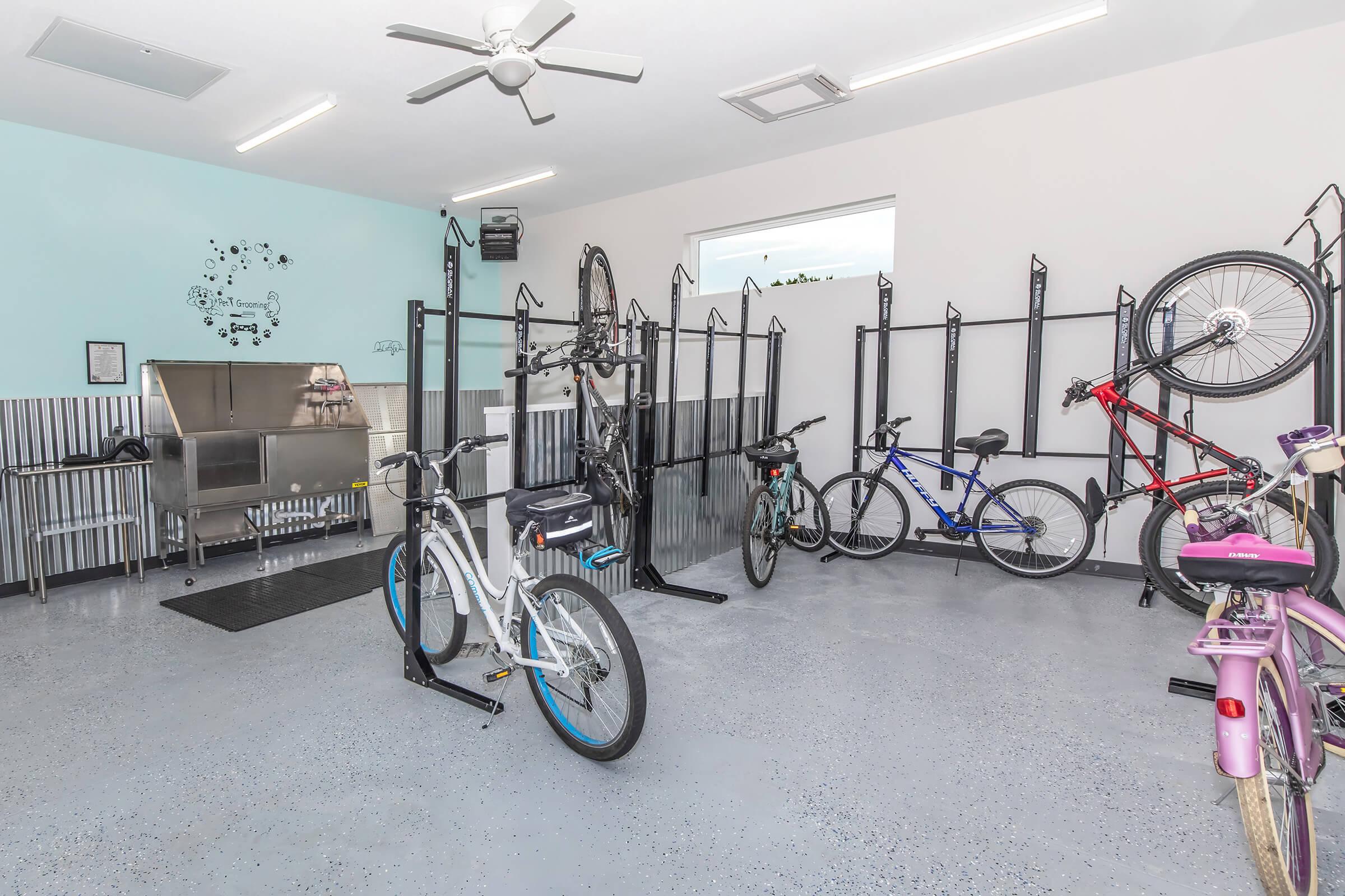 A spacious bike storage area with various bicycles parked on metal racks. The room features a light blue wall, a ceiling fan, and a polished concrete floor. There are also bicycle repair tools and equipment visible, creating an organized and functional environment for bike maintenance and storage.