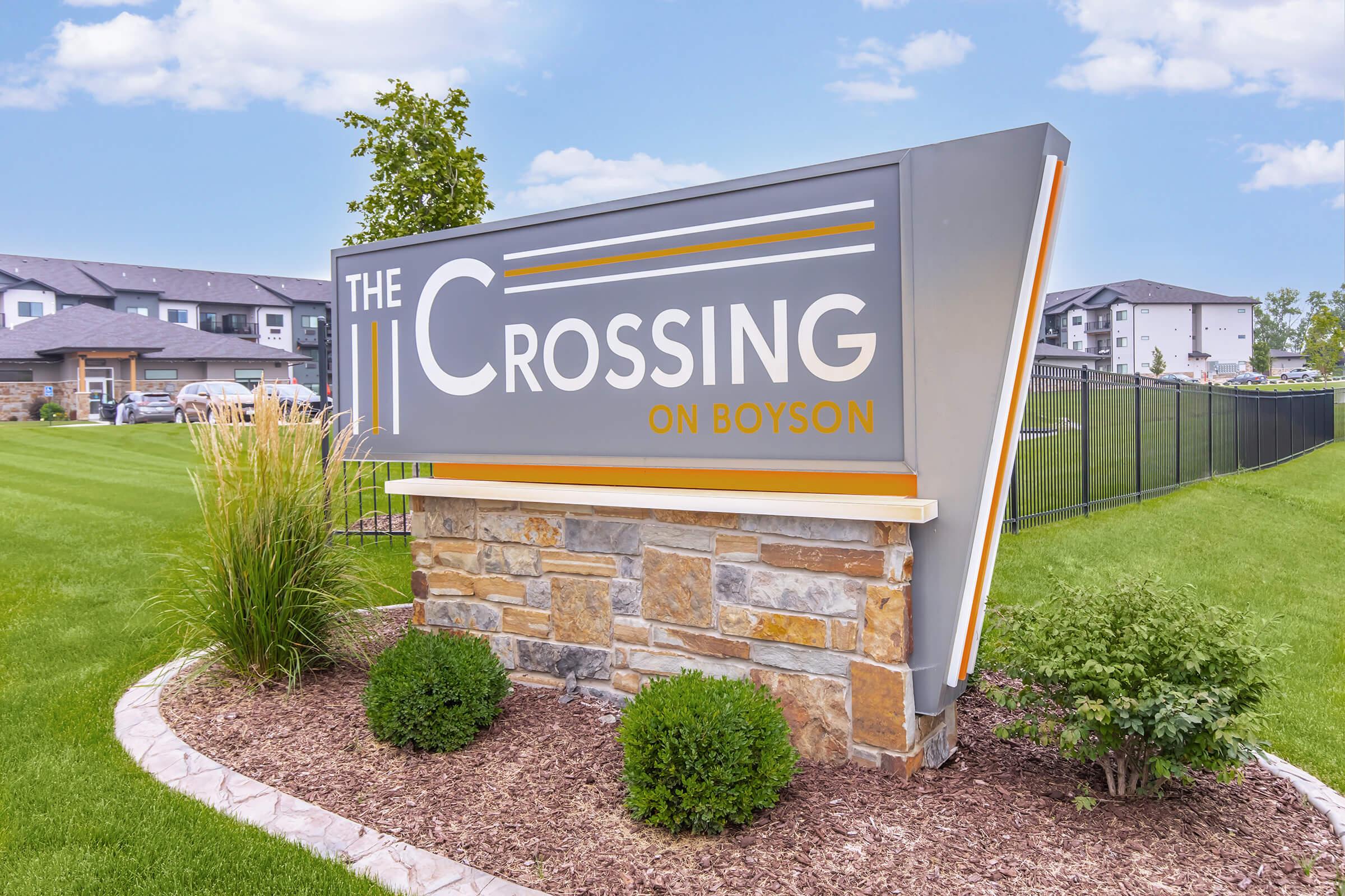 Sign for "The Crossing on Boyson," featuring modern design with gray and orange colors. Surrounded by landscaped grass and shrubbery, the sign prominently displays the name and is set against a clear blue sky, conveying an inviting atmosphere for visitors.