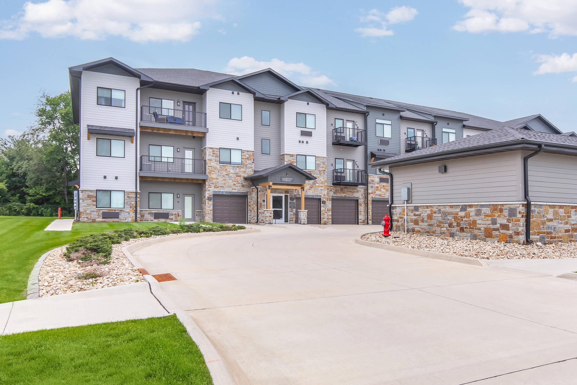 Modern multi-unit residential building with a combination of stone and siding exterior. The structure features balconies and garages, surrounded by a well-maintained lawn and landscaping. A paved driveway leads into the complex, creating a welcoming entrance.
