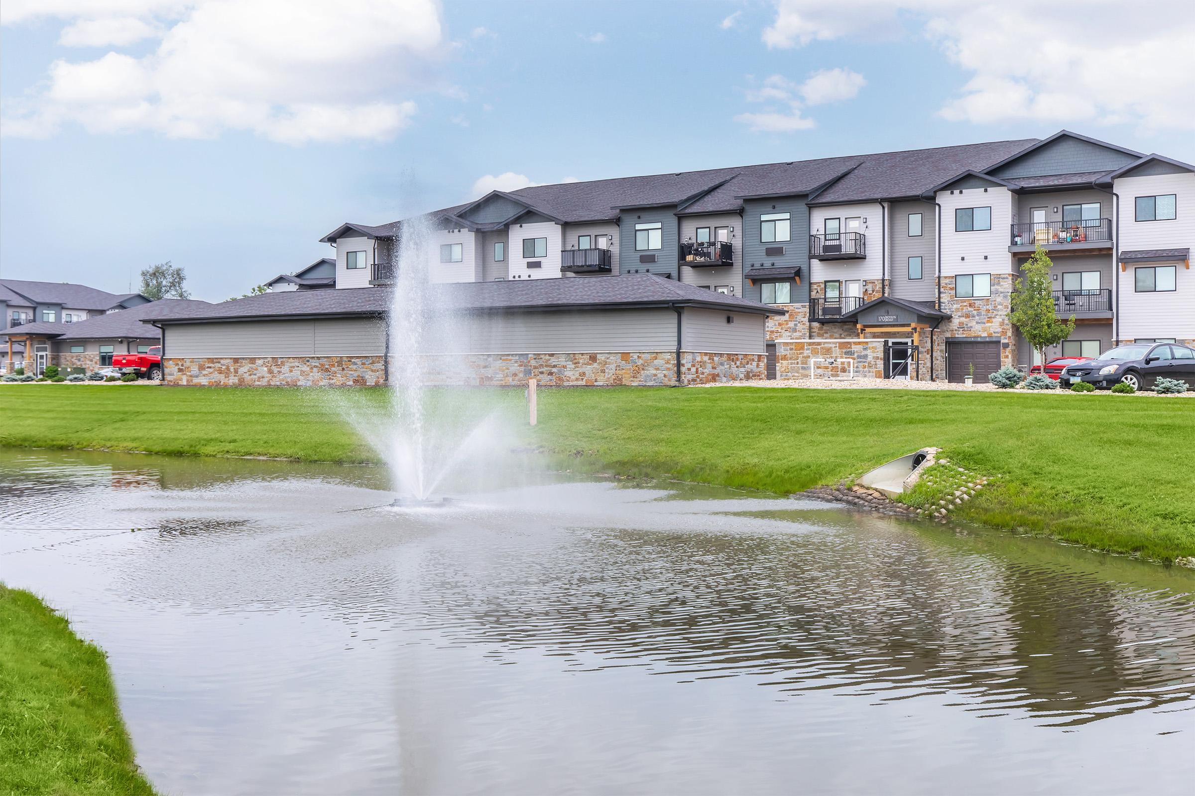 A serene residential area featuring an artificial pond with a fountain in the foreground. Behind the pond, there are modern multi-unit apartment buildings with balconies. Lush green grass surrounds the pond, and a few parked cars are visible nearby under a partly cloudy sky.