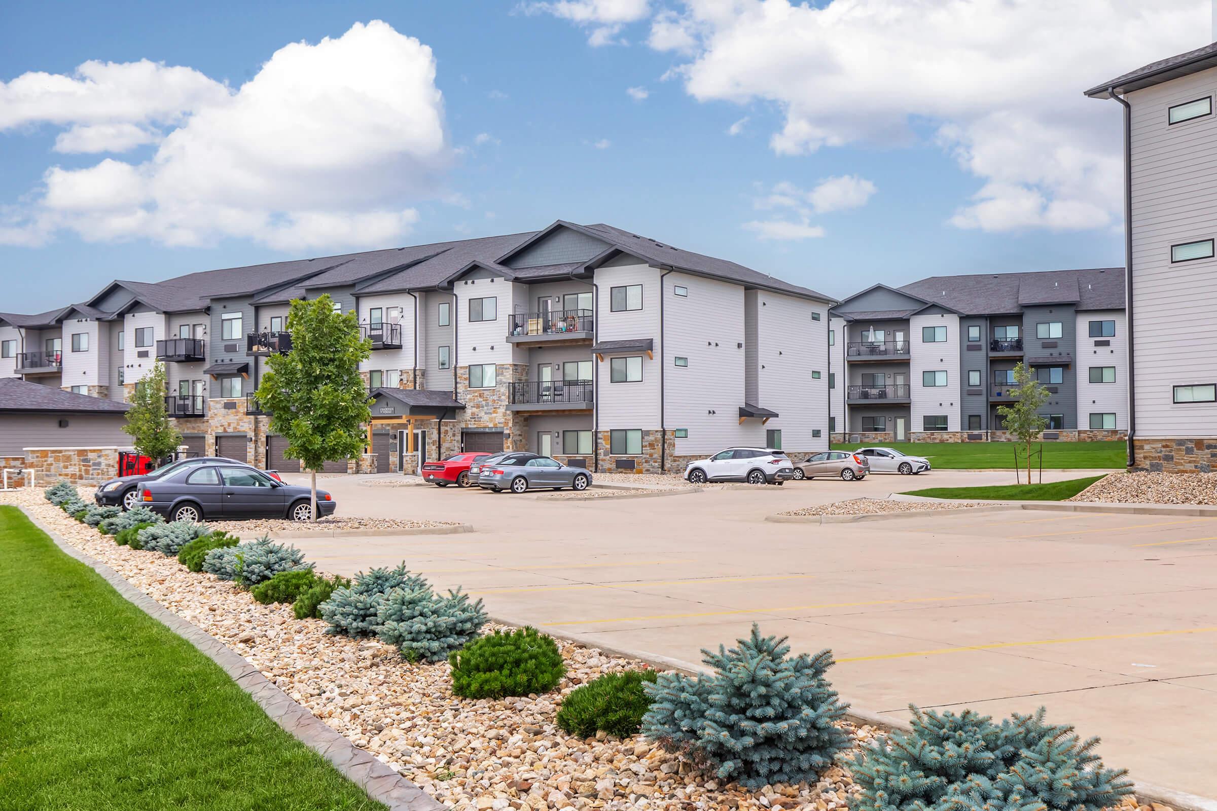 A modern apartment complex featuring several buildings with balconies, set against a backdrop of blue sky and clouds. The well-maintained landscape includes grassy areas and decorative shrubs, with a parking lot visible that has multiple parked cars.
