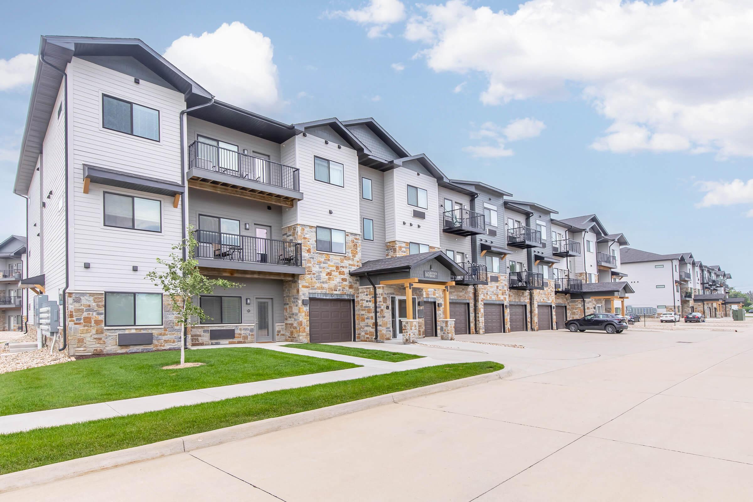 A row of modern multi-family residential buildings with stone accents and balconies. The buildings are two to three stories high and feature garages at the ground level. A well-maintained lawn and paved area are in the foreground, with a clear blue sky and fluffy white clouds in the background.