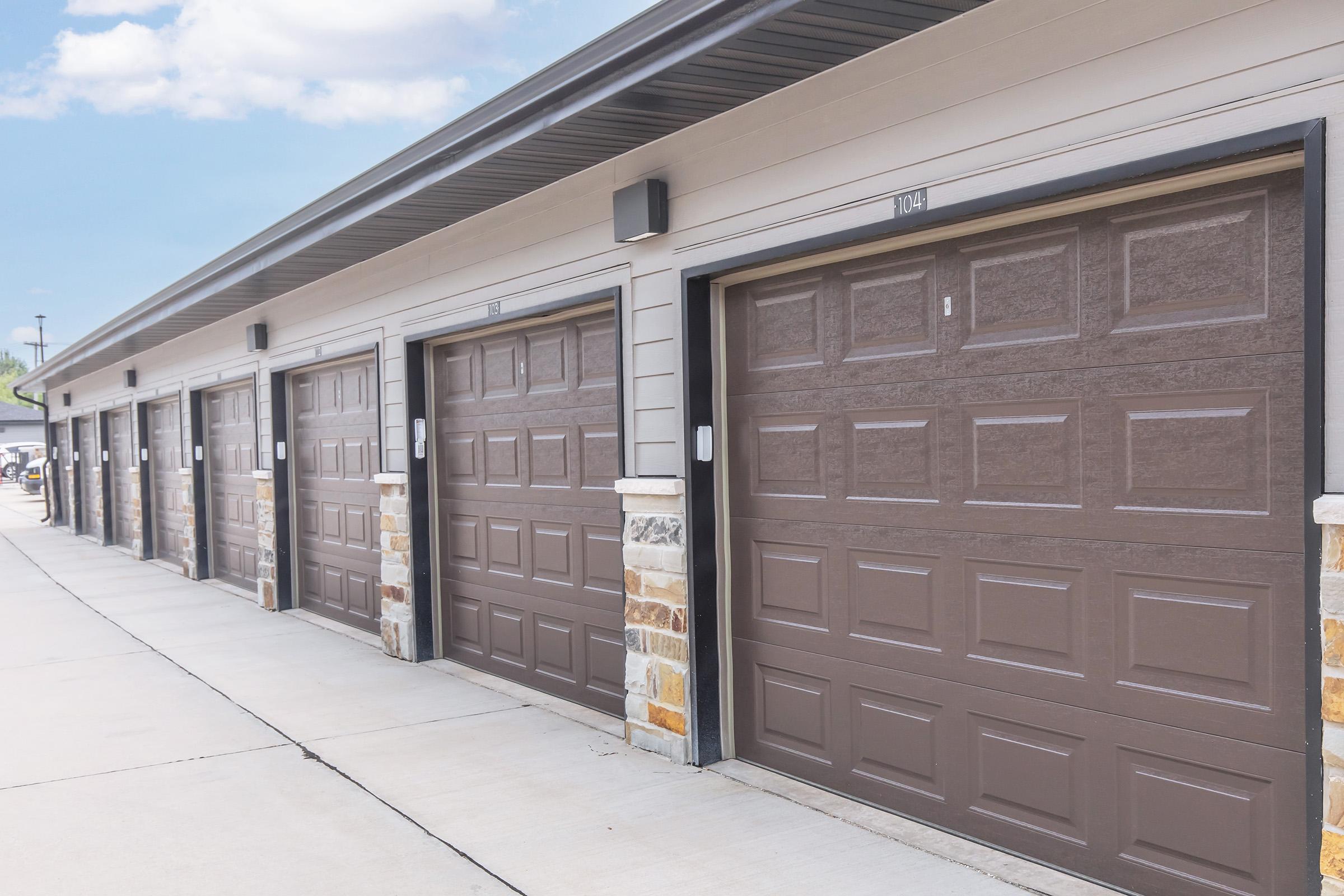Row of beige garage doors with brown panels, set against a light gray building. Each garage door has a number visible on the top. The area is paved with concrete and features stone accents near the base of the building. The sky is clear with a few clouds.
