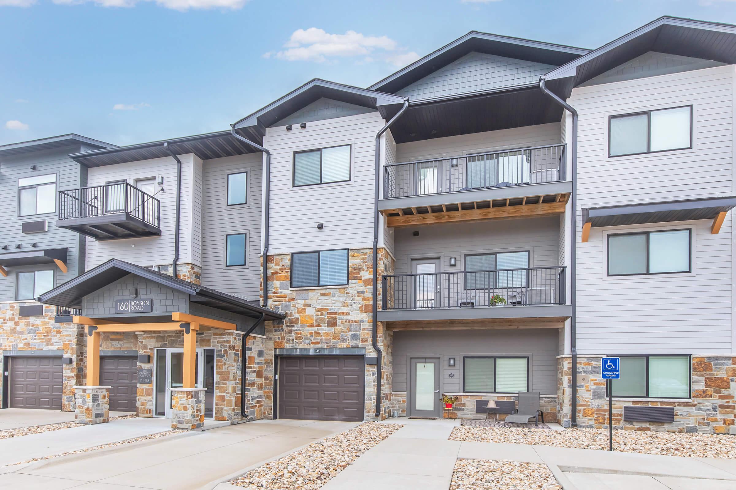 A modern multi-unit apartment building featuring a combination of gray siding and stone accents. The building has balconies, large windows, and garage doors. The entrance is marked with signage and there is a small landscaped area in front. The sky above is clear with a few clouds.