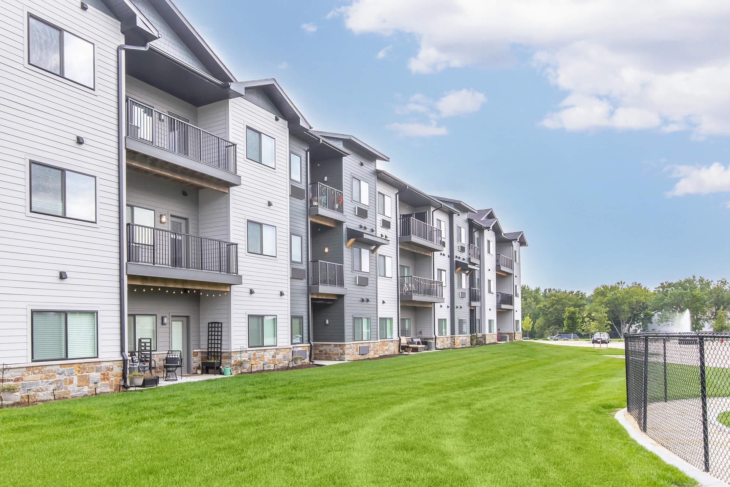 A row of modern apartment buildings with multiple balconies and stone accents. The buildings are set against a clear blue sky and feature green grass in the foreground. A fence borders the area, suggesting a well-maintained outdoor space for residents.
