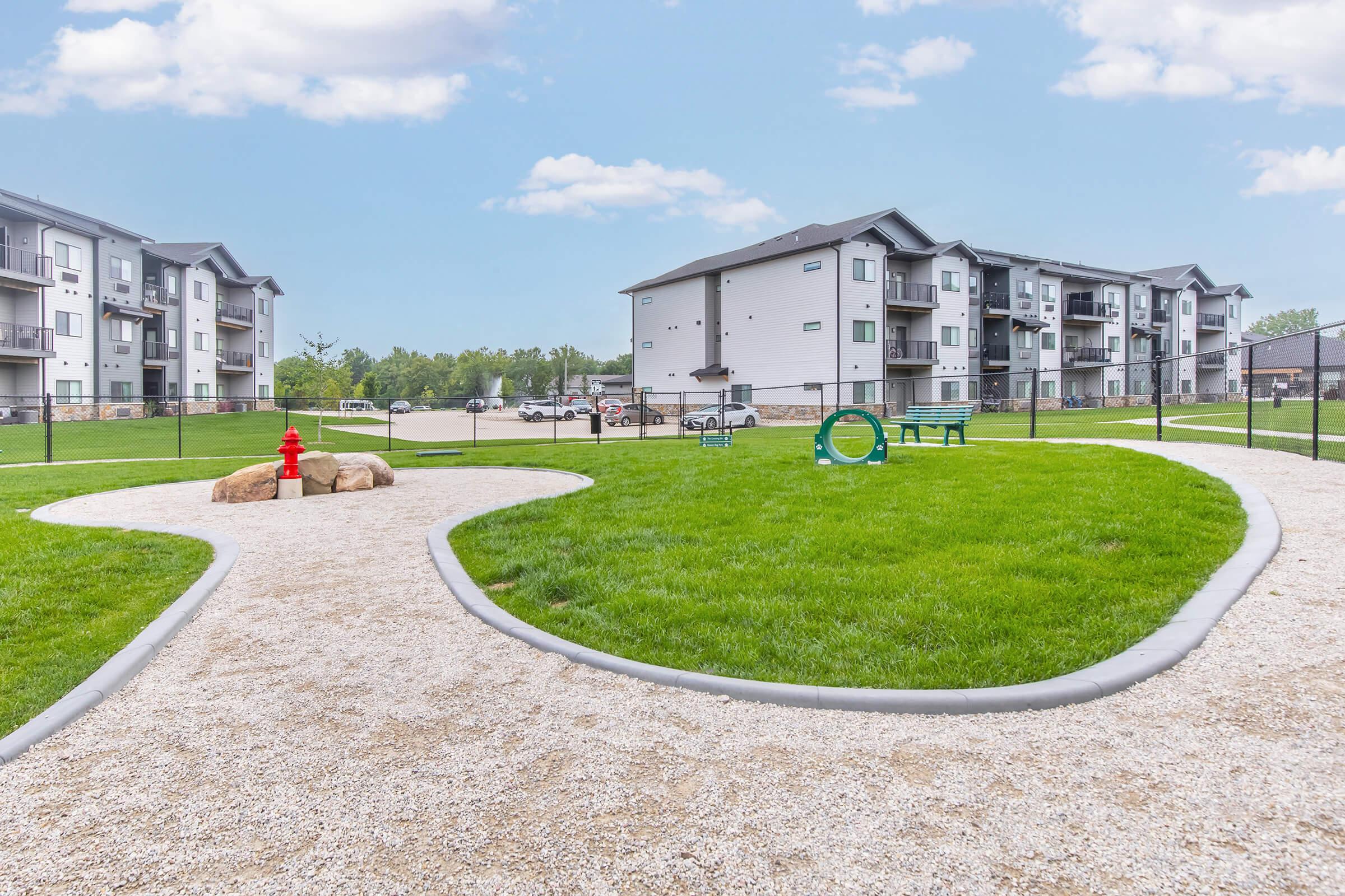 A well-maintained dog park featuring a gravel pathway winding through lush green grass. There are rocks for seating, a fire hydrant, and a green agility tunnel. In the background, modern apartment buildings are visible under a clear blue sky. The area is enclosed with a fence, creating a safe space for pets.