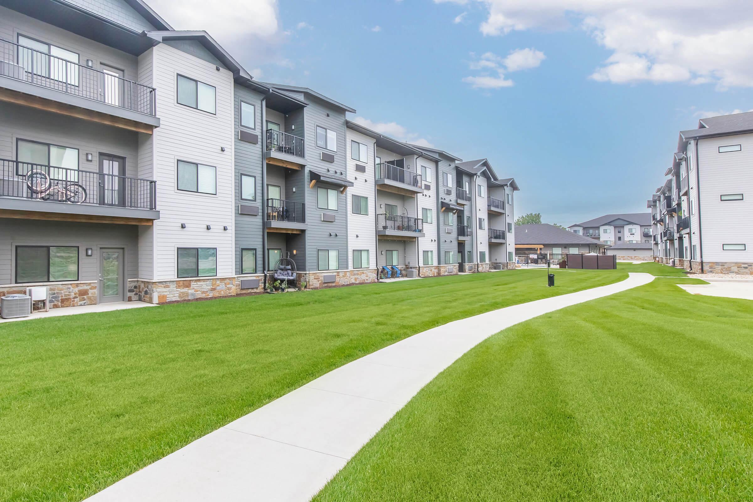 A well-maintained apartment complex with multiple three-story buildings. The buildings feature balconies and large windows, set against a backdrop of a clear blue sky. A lush green lawn and a paved walking path wind through the yard, adding to the inviting outdoor space.