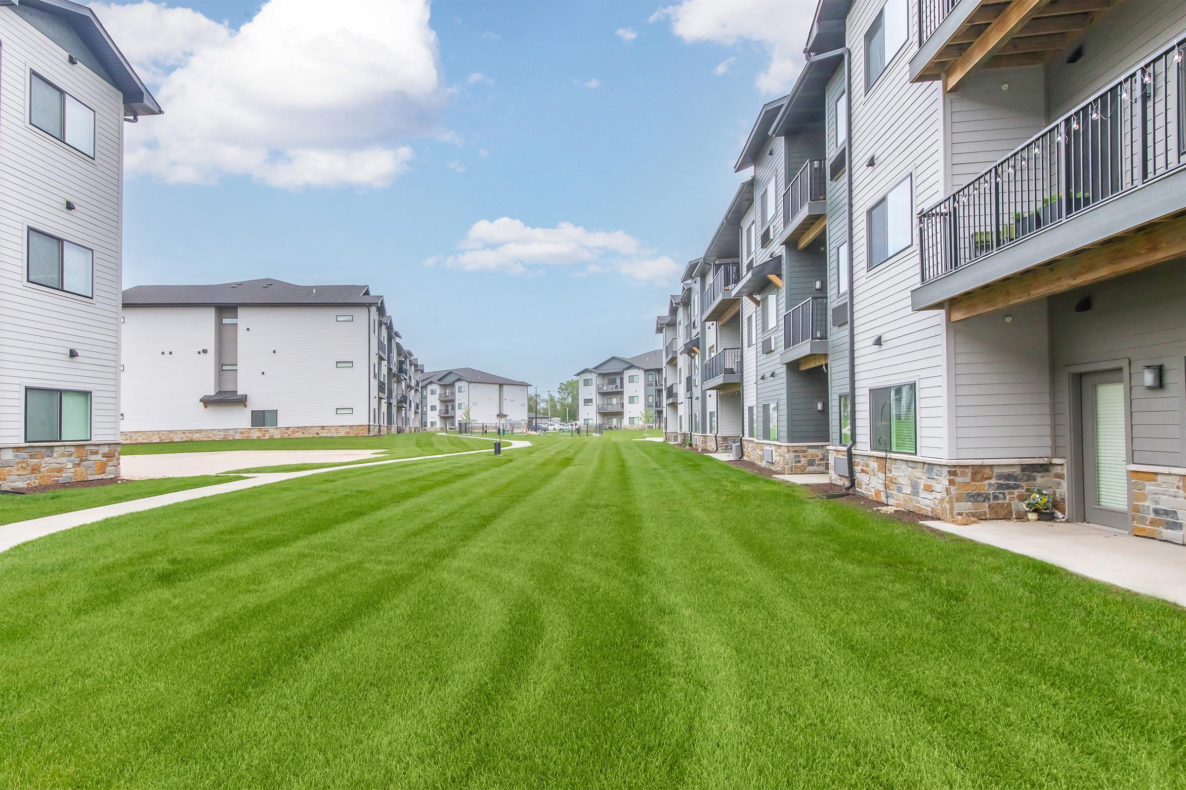 A view of a landscaped area between modern apartment buildings, featuring green grass and walkways. The buildings have a contemporary design with balconies and stone accents. The sky is bright with scattered clouds, creating a sunny atmosphere.