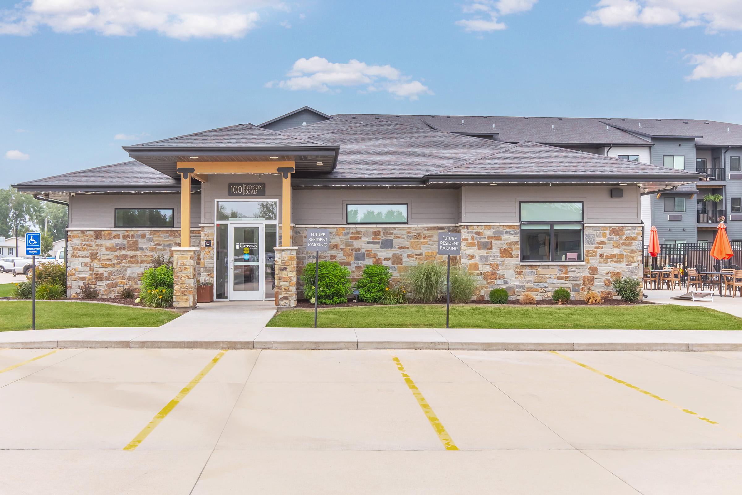 A modern building with a stone facade and a welcoming entrance. The structure features large windows and a sloped roof, surrounded by well-maintained landscaping and a paved parking area. In the background, there are tables with umbrellas, suggesting an outdoor dining or relaxation space.
