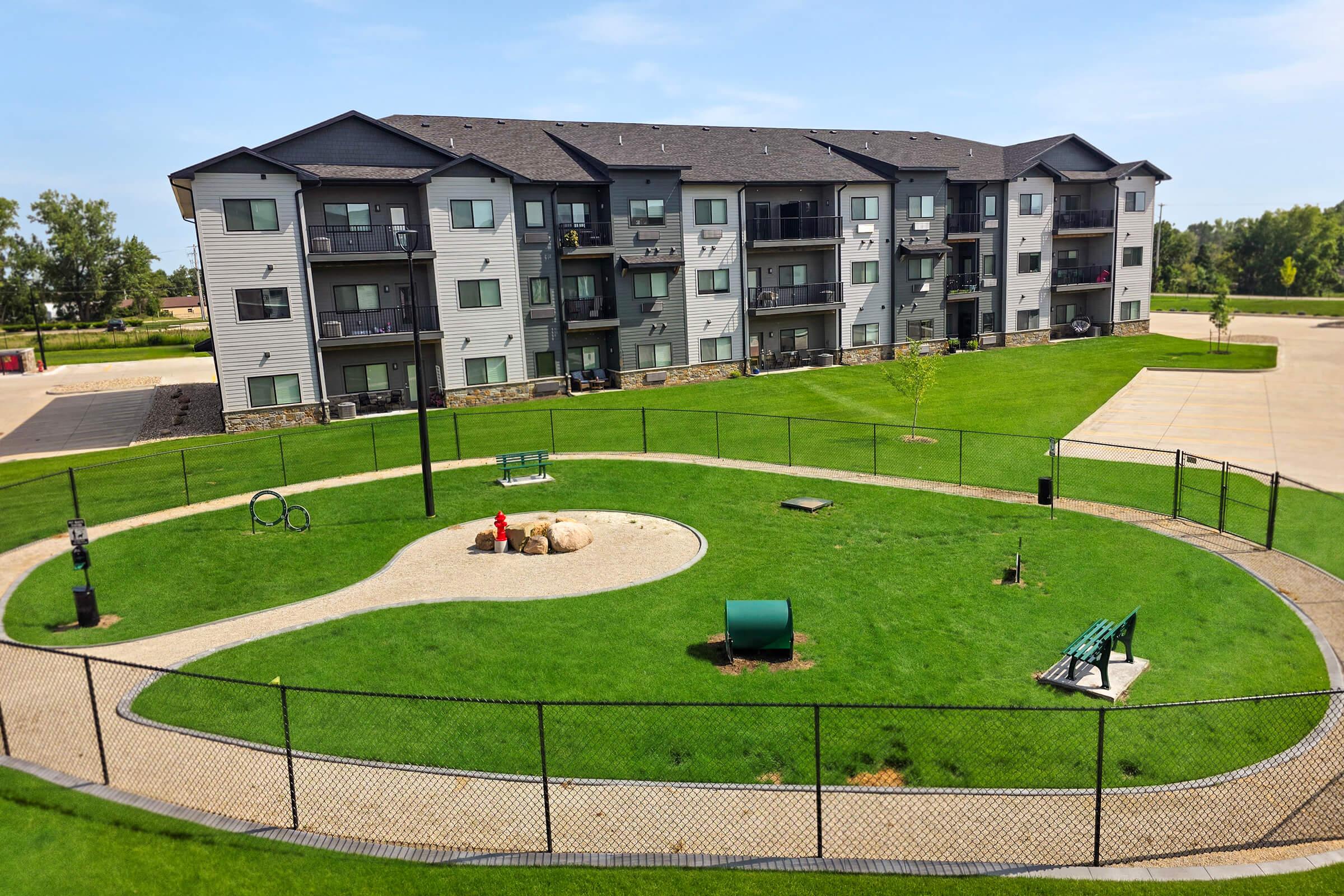 A well-maintained green area featuring a fenced dog park with various amenities, including rocks, a bench, and play equipment. In the background, a modern apartment building with balconies is visible under a clear blue sky.