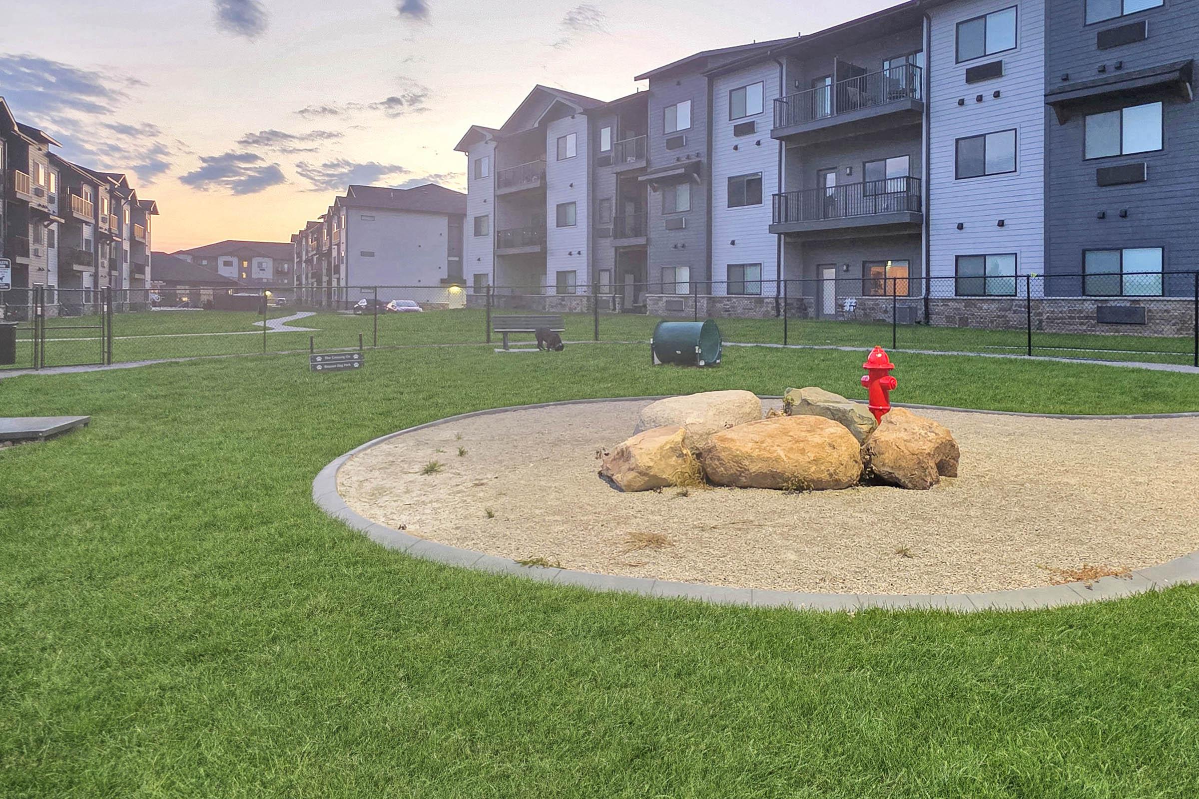 A grassy area with a circular gravel patch featuring large rocks at the center. A red fire hydrant stands prominently among the rocks. In the background, modern apartment buildings are visible, with benches and a waste bin nearby. The sky shows a warm color palette indicative of sunset.