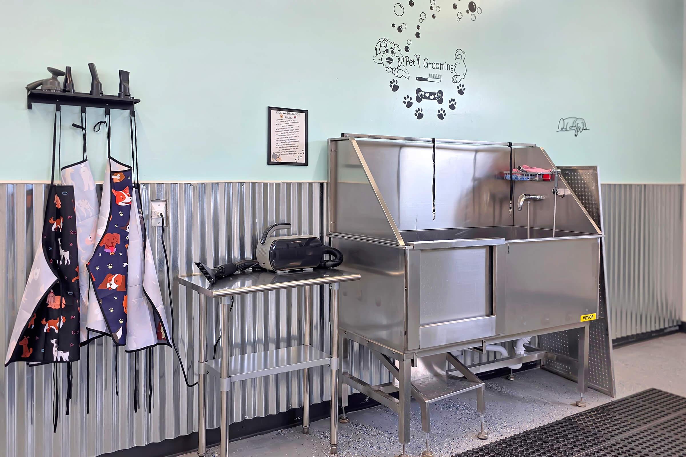 A clean pet grooming area featuring a stainless steel wash station, a drying machine, and grooming aprons hanging on a wall. The walls are light blue with paw print decorations, creating a welcoming atmosphere for pets.