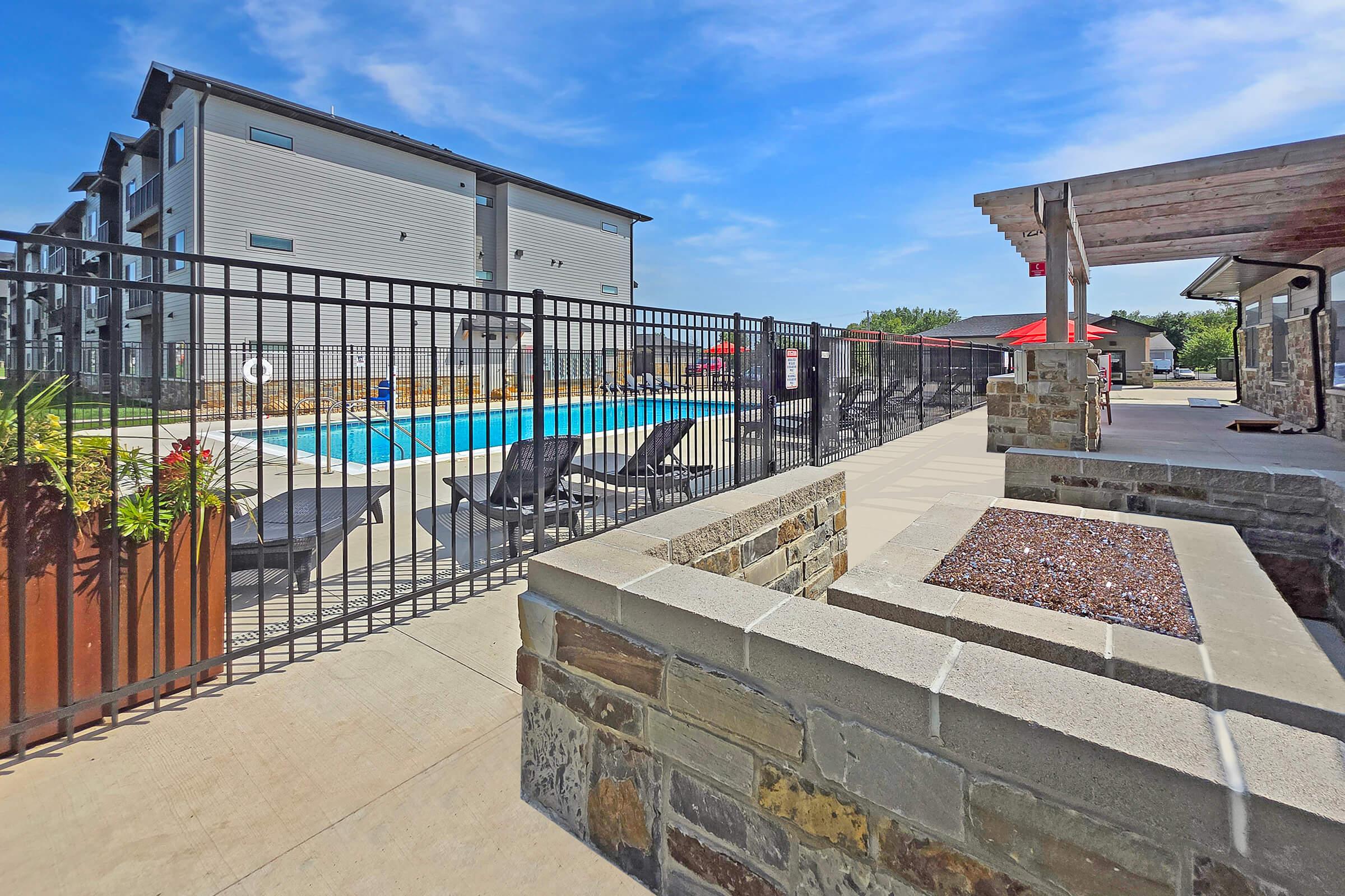 A well-maintained outdoor pool area surrounded by lounge chairs and a decorative stone wall. In the background, modern apartment buildings are visible. The scene is bright with clear blue skies and features a safety fence around the pool, creating an inviting recreational space.