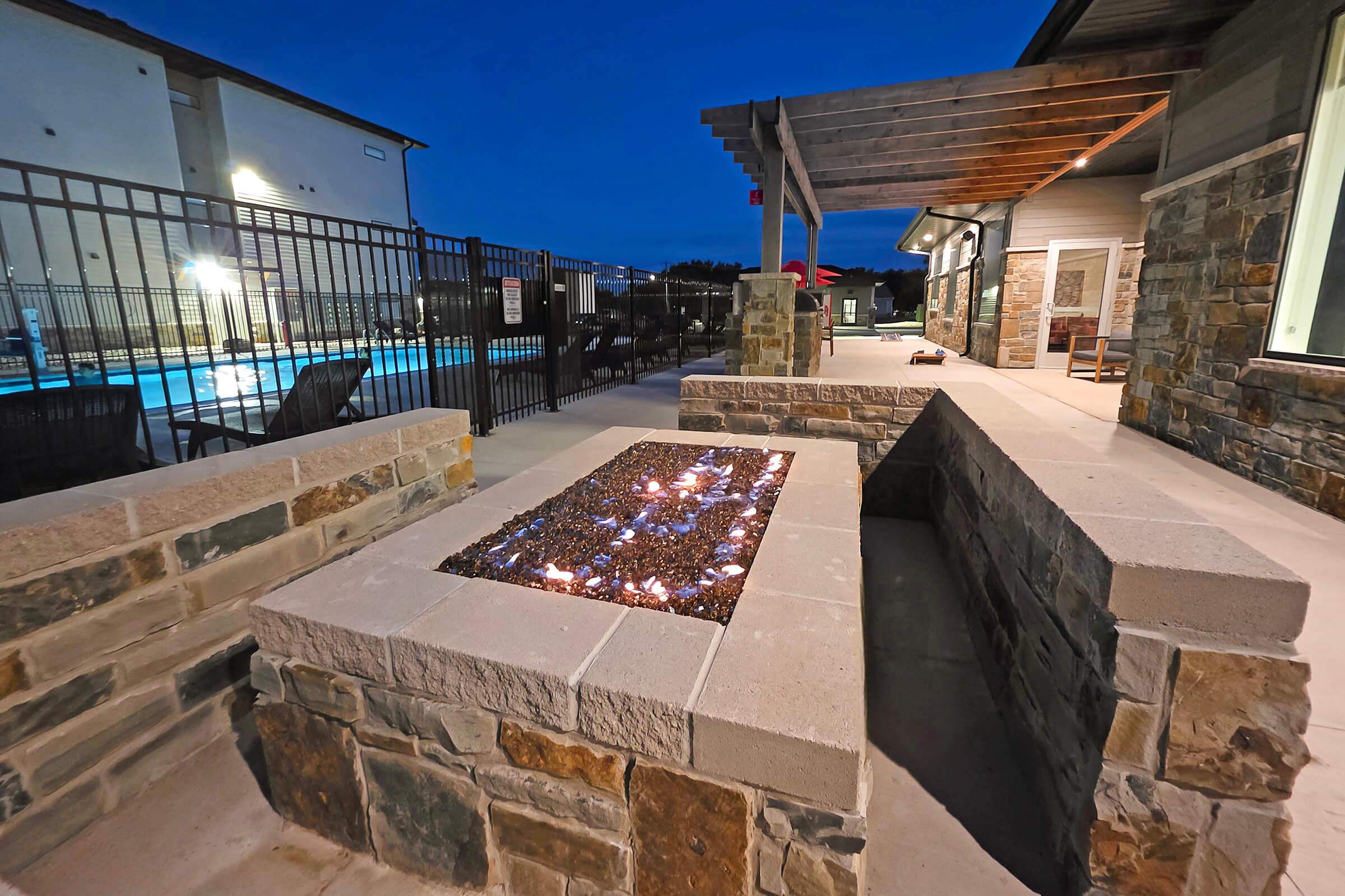 Stone fire pit surrounded by a concrete seating area, illuminated in the evening. In the background, there is a fenced swimming pool area with lounge chairs, and a partially covered patio. The sky is darkening, adding a cozy ambiance to the setting.