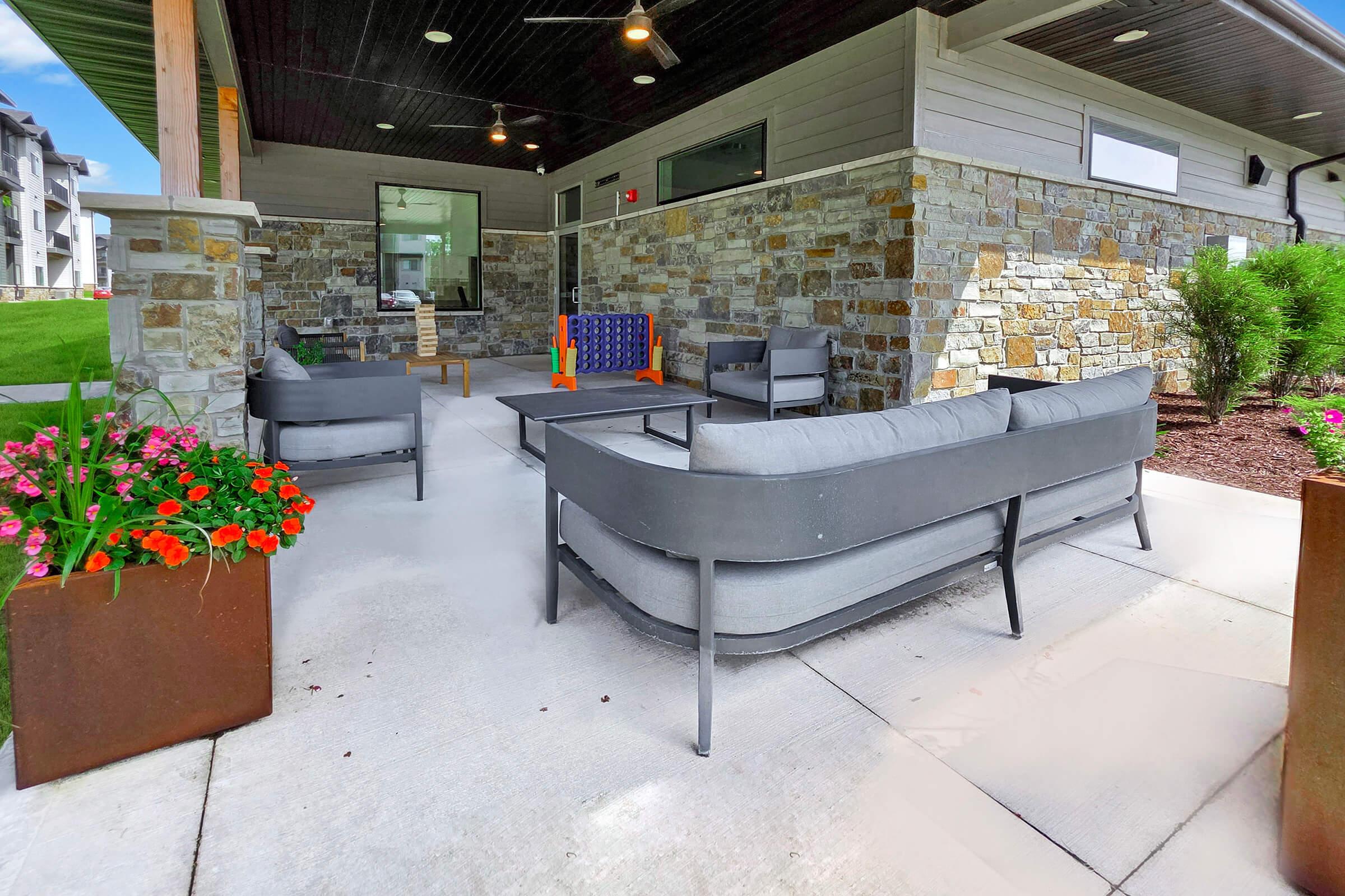 Outdoor patio area featuring grey seating furniture arranged around a small table, surrounded by colorful flower planters and modern stone walls. Natural light fills the space, enhancing the inviting atmosphere. Ideal for relaxation or social gatherings.