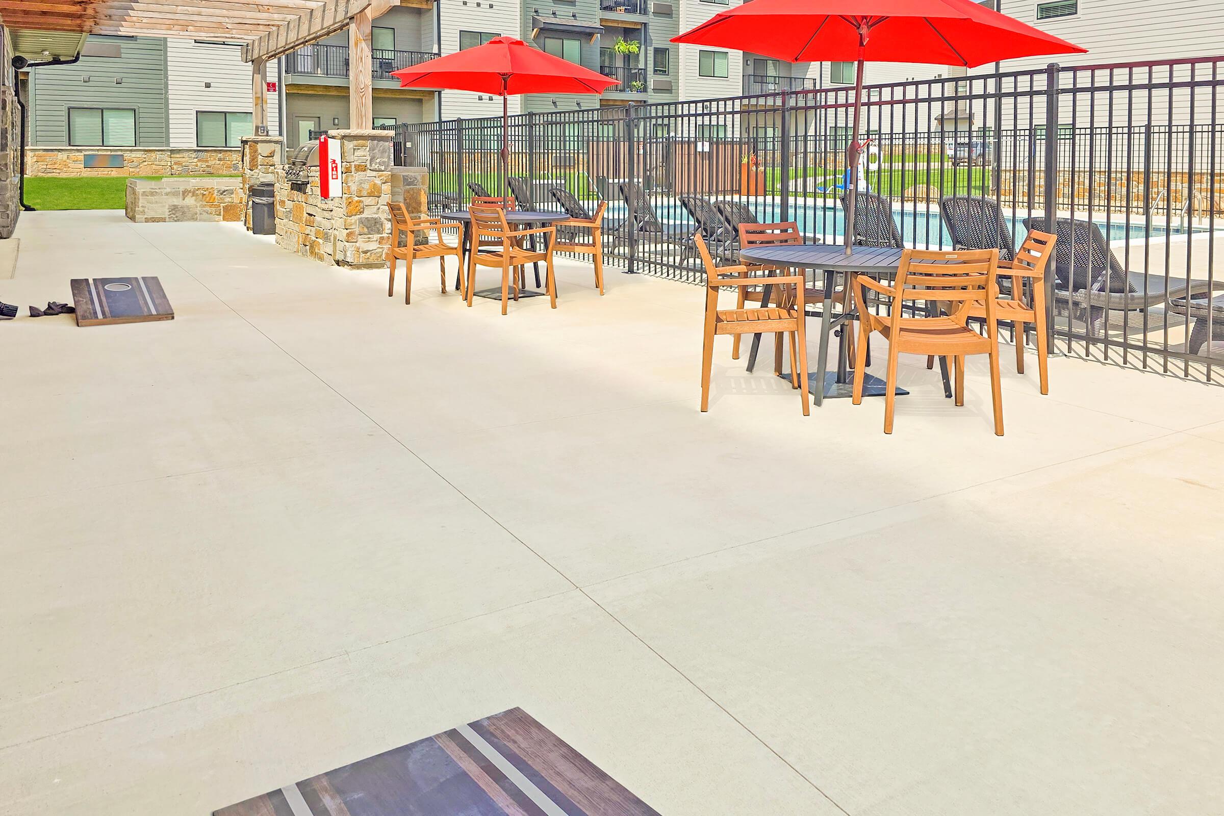 Outdoor patio area featuring several black tables and chairs, each with bright red umbrellas. The setting includes a swimming pool in the background and is surrounded by a decorative stone wall. The space is well-lit and inviting, ideal for relaxation or dining.