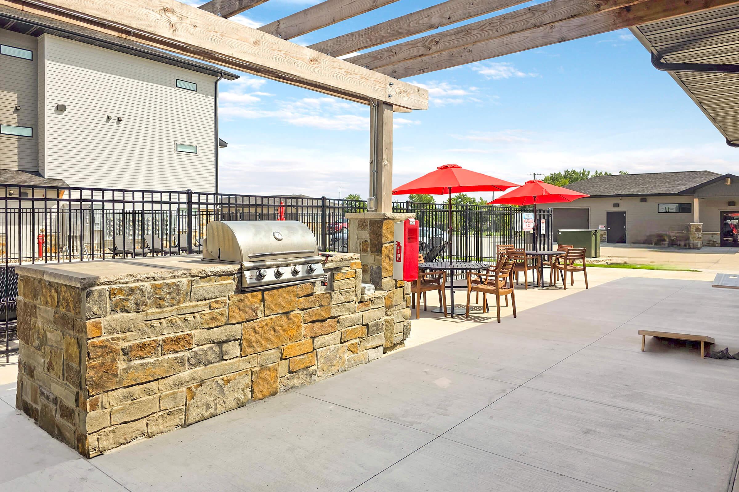 Grilling area with a stone grill under a wooden pergola. Surrounding it are a few metal chairs and tables with red umbrellas. In the background, residential buildings and a fence are visible, along with a clear blue sky.