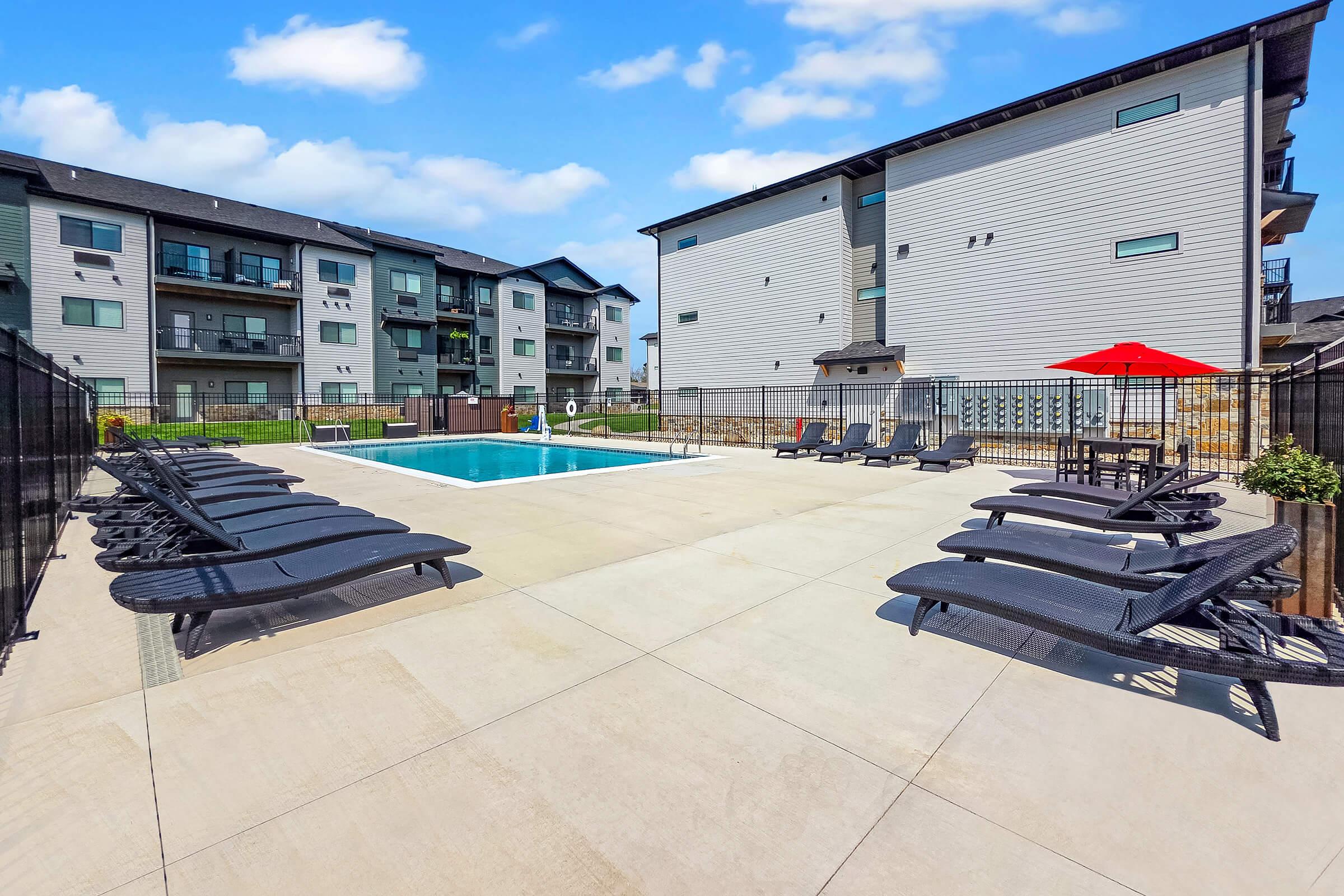 A modern swimming pool area surrounded by lounge chairs, with a red umbrella providing shade. In the background, there are multi-story apartment buildings. The pool area is fenced, and the concrete surface is clean and well-maintained, reflecting a bright, sunny day.