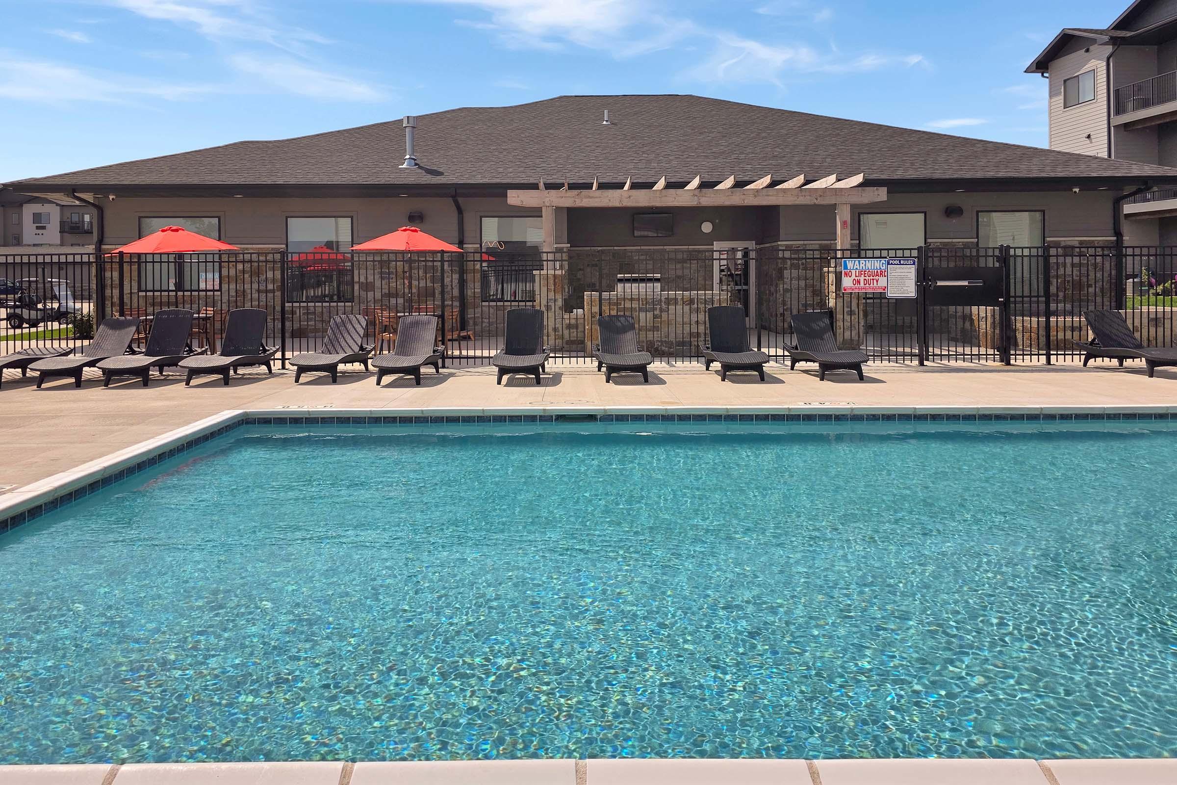 Swimming pool with clear blue water in the foreground, surrounded by lounge chairs. A shaded area with red umbrellas is visible in the background near a building with a brown roof and stone accents. The area is enclosed with a black fence, creating an inviting outdoor space.