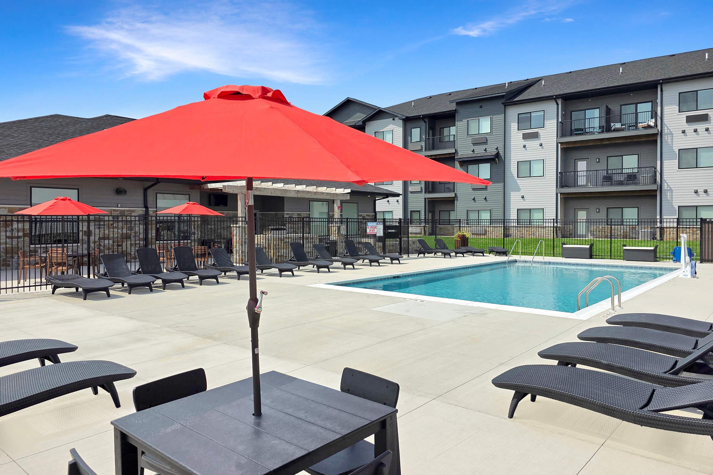 A swimming pool area with a clear blue sky overhead. The pool is surrounded by black lounge chairs and tall red umbrellas, providing shade. In the background, modern apartment buildings are visible. The setting is inviting, perfect for relaxation and leisure on a sunny day.