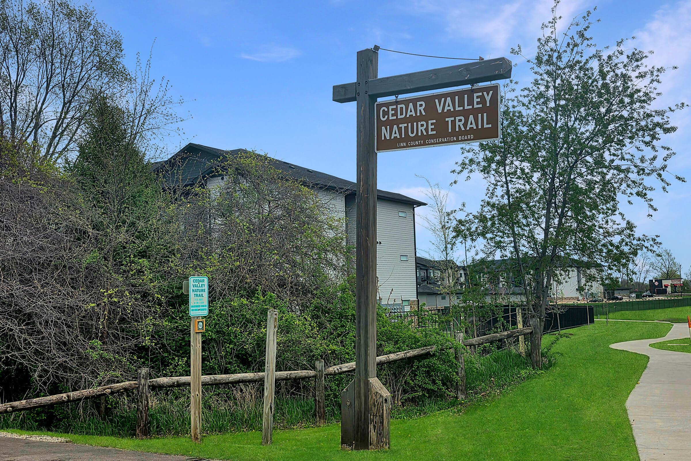 A wooden sign reading "Cedar Valley Nature Trail" stands at the entrance of a green pathway. Nearby, a fenced area and a few trees frame the path, with a modern building partially visible in the background. The scene is peaceful, suggesting a connection with nature.