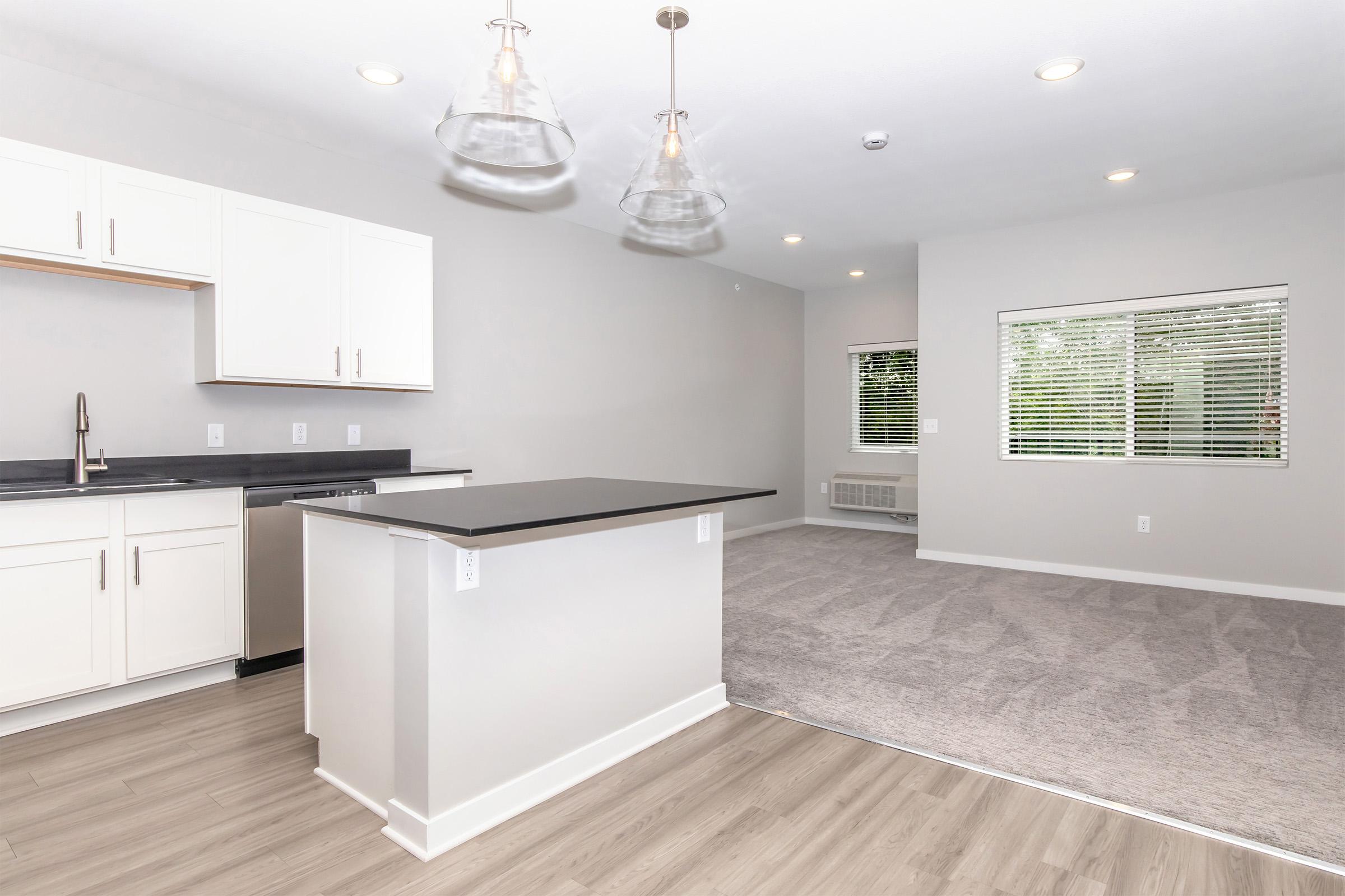 A modern open-concept kitchen and living area with light gray walls and wood-style flooring. The kitchen features white cabinetry, a dark countertop, and stainless steel appliances. Bright pendant lights hang above the kitchen island. The living space has large windows allowing natural light to enter, and a cozy carpeted area is visible.