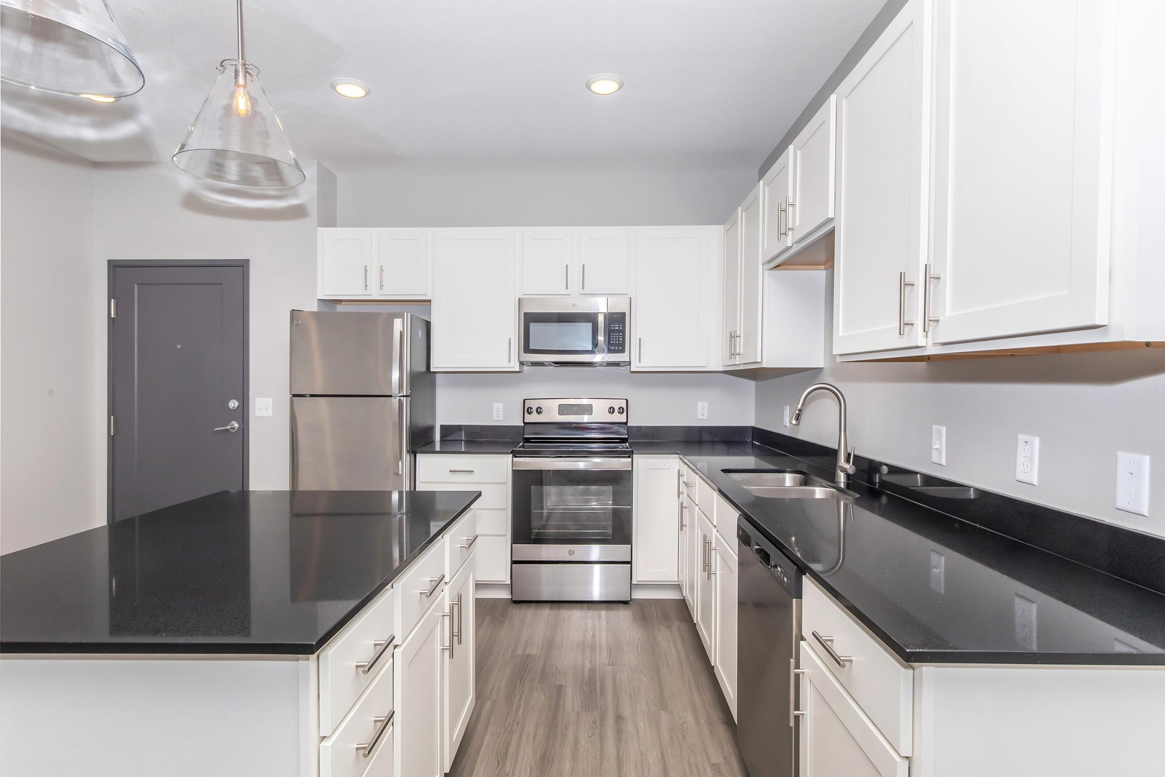 Modern kitchen featuring white cabinetry, stainless steel appliances including a refrigerator, microwave, and oven. The countertops are dark granite, complemented by a large island for food preparation. The space is well-lit with recessed lighting and pendant fixtures, creating a bright and inviting atmosphere.
