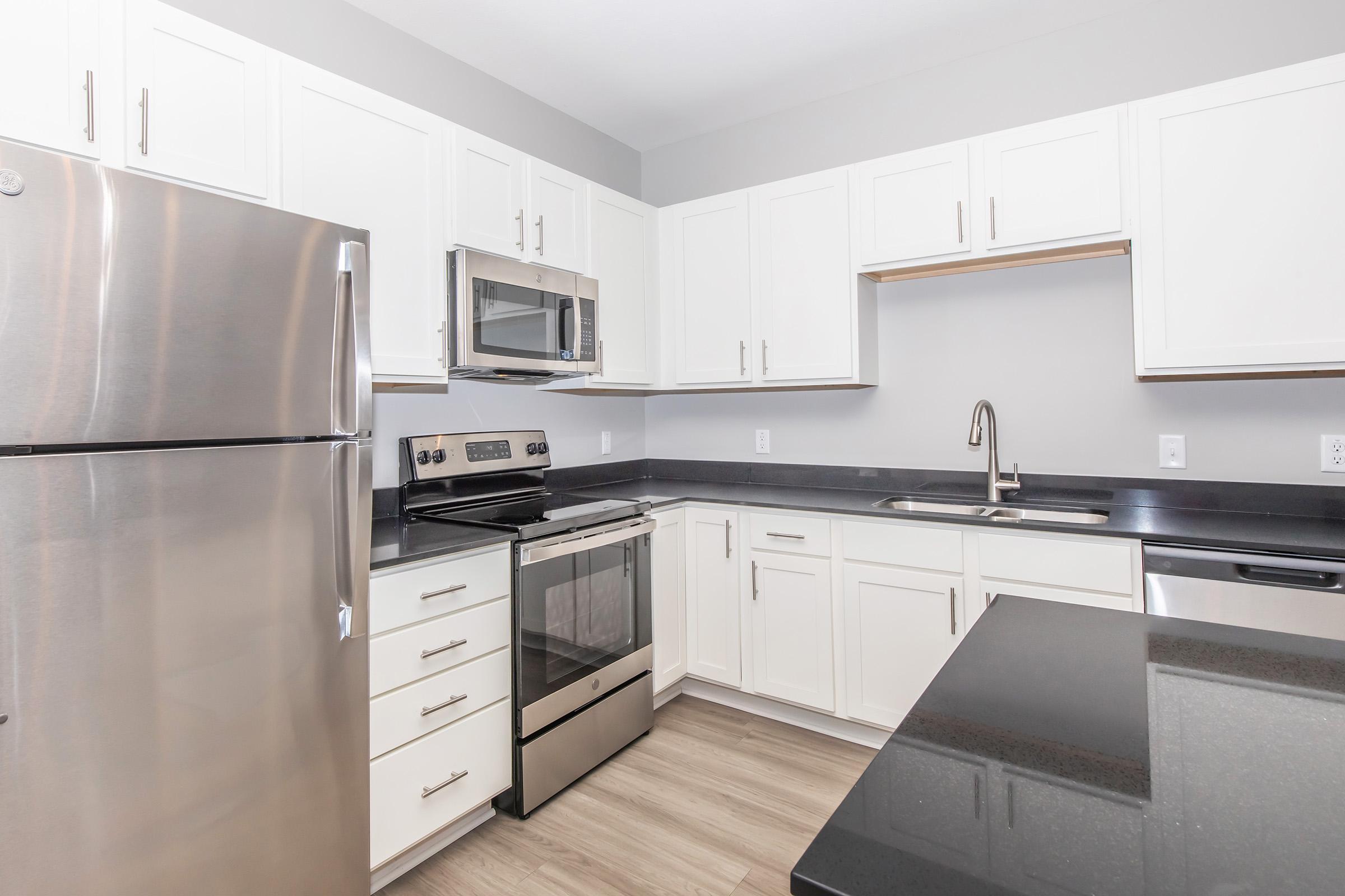 Modern kitchen featuring stainless steel appliances, including a refrigerator, oven, and microwave. The cabinetry is white, complemented by a dark countertop. A single-basin sink is installed, and there's a spacious island in the center, enhancing the kitchen's functionality and style. Floors are light-colored wood.