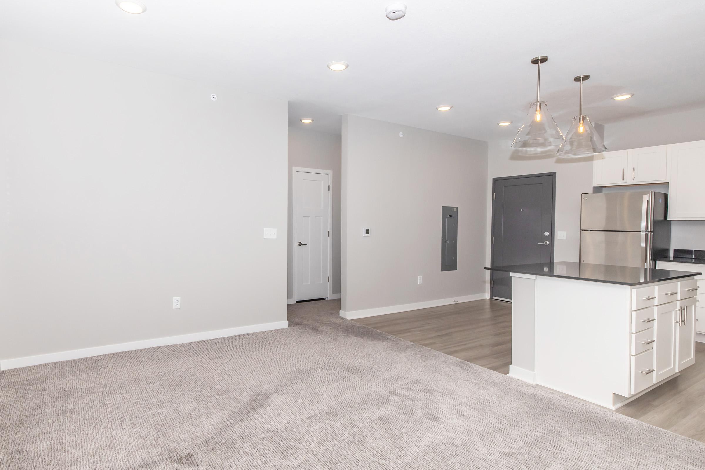 A spacious, modern living area with light gray walls and a carpeted floor. The room features a kitchen with white cabinetry, stainless steel appliances, and a dark countertop. Three pendant lights hang above the kitchen island, and there's an open doorway leading to another room.