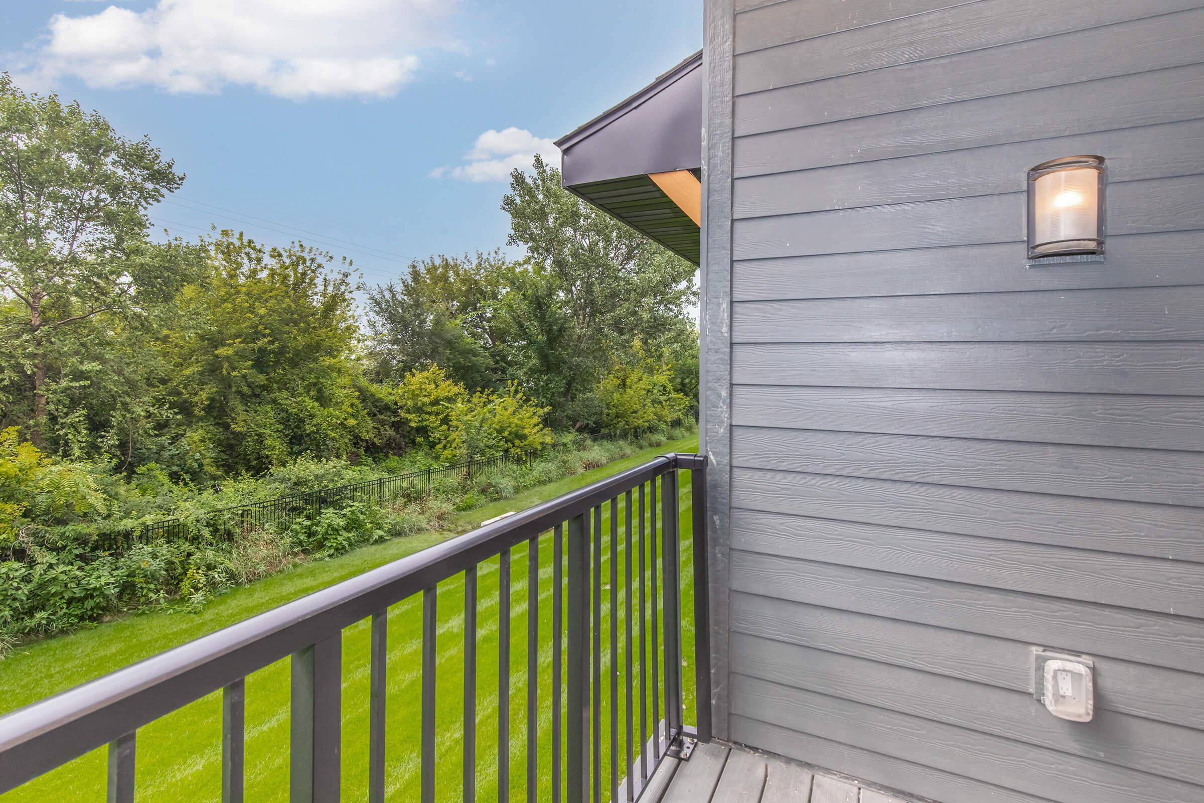 View from a balcony showing a modern wooden railing leading to a lush green lawn and tree line in the background, under a partly cloudy sky. A light fixture is attached to the side of the building, enhancing the outdoor space.