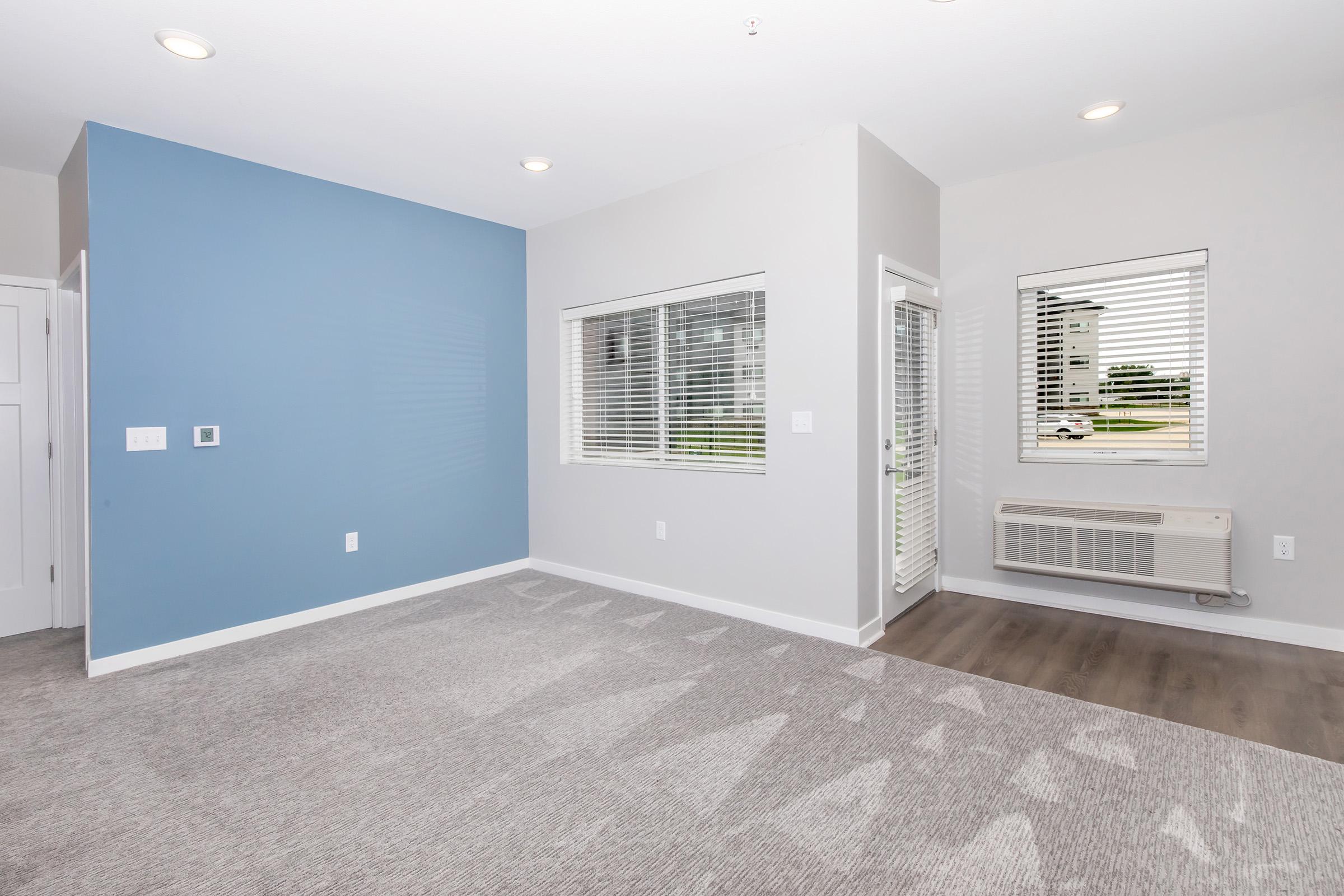 Spacious interior of a modern room featuring light gray carpet, a blue accent wall, and large windows with blinds. The room includes an air conditioning unit and door leading outside, with neutral-colored walls giving a bright and inviting atmosphere.