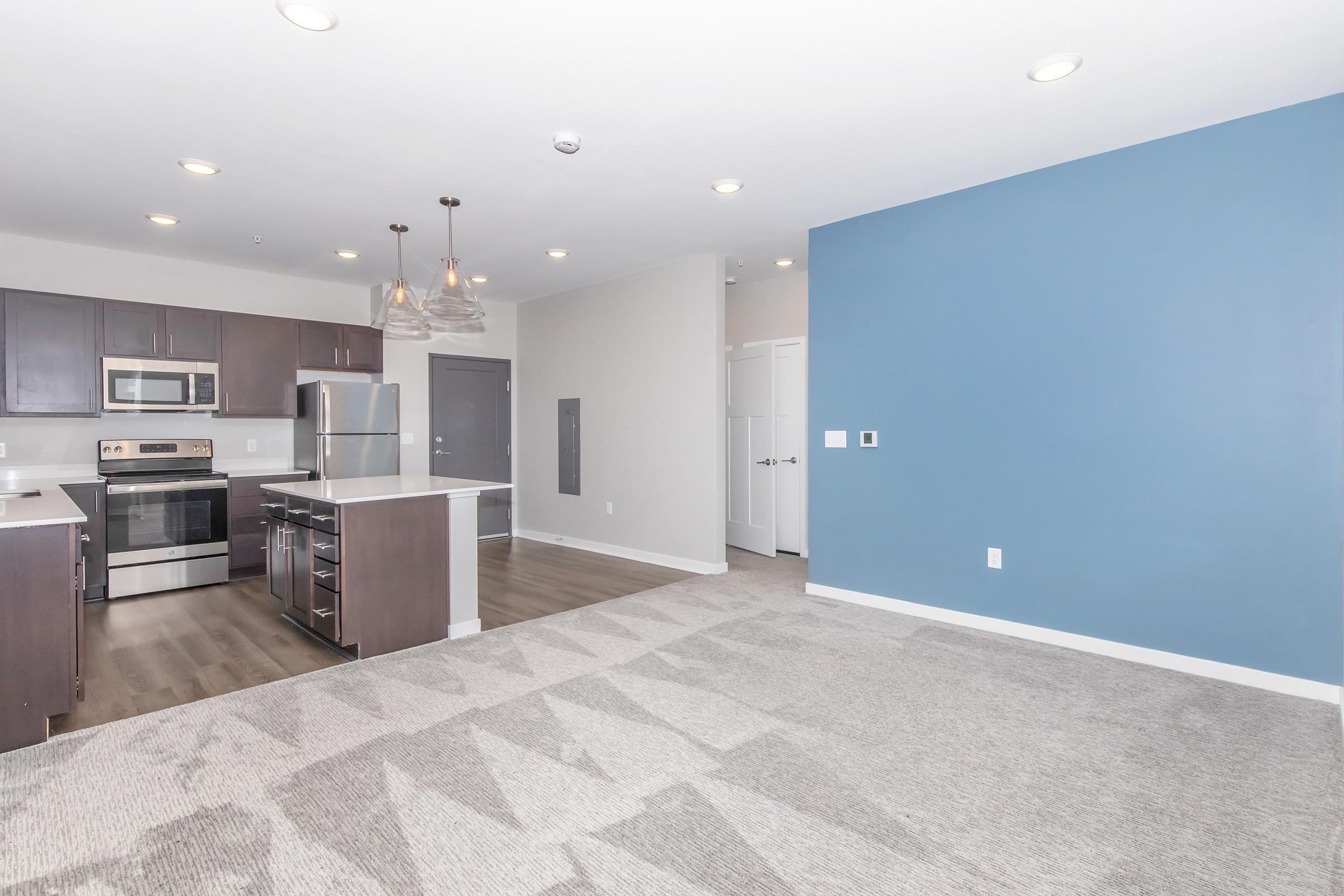 Modern apartment interior featuring a kitchen with stainless steel appliances, a large island, and dark wood cabinetry. The living area includes light gray carpeting and a blue accent wall, with ample natural light. There are white doors leading to additional rooms in the background.